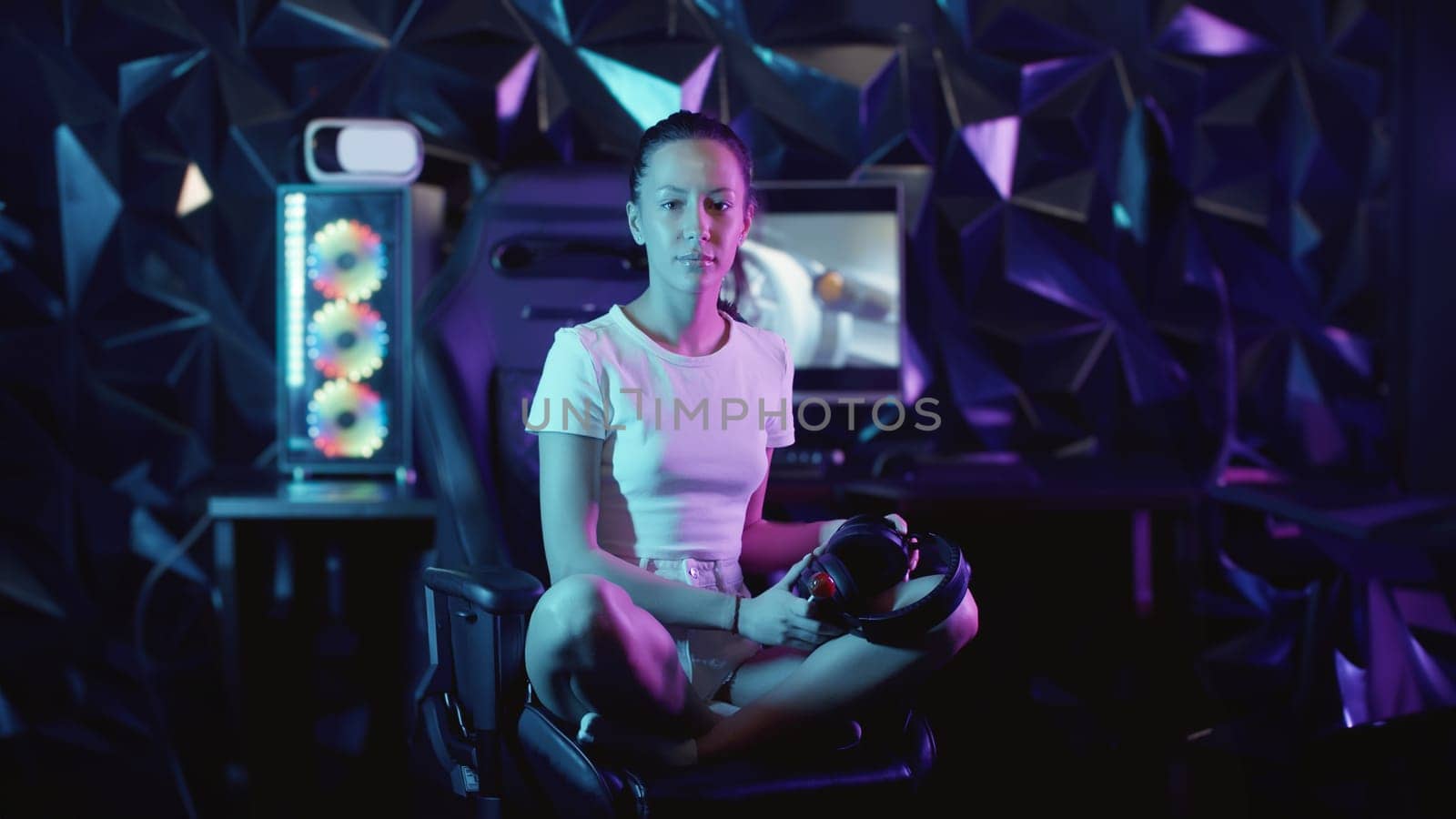 Girl gamer sits on the chair in the gaming club and putting on her headphones - sucking lollipop and looking in the camera, portrait