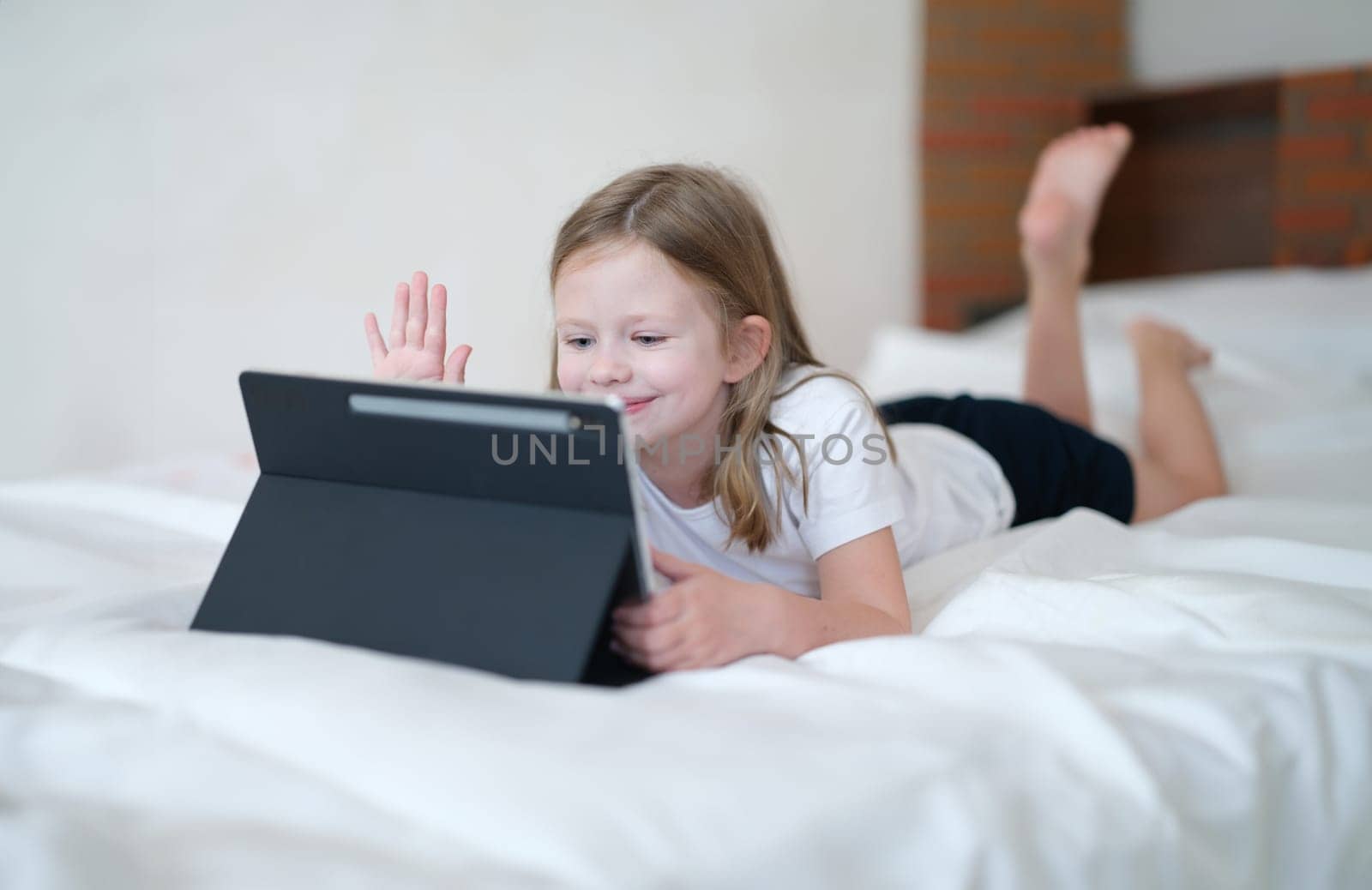 Little girl lying on bed and waving her hand at screen of tablet by kuprevich