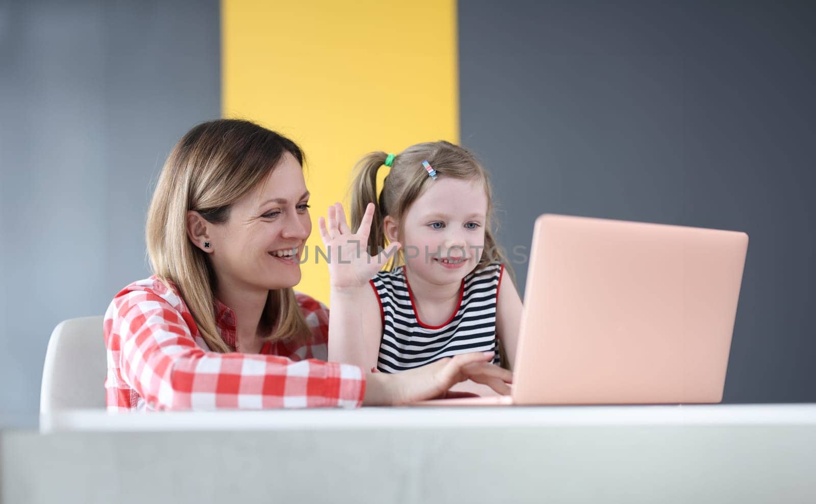 Mother and little daughter are sitting in front of laptop and waving by kuprevich