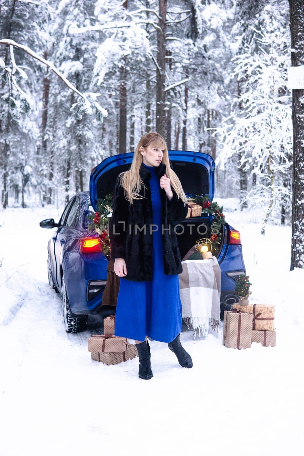 Woman in winter snowy forest in blue dress next to blue car decorated with Christmas decor. Christmas and winter holidays concept