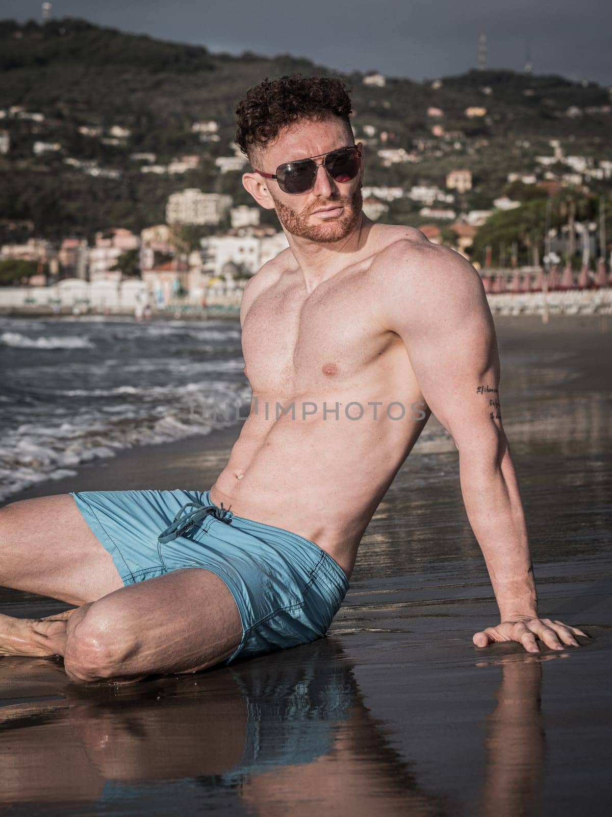 A shirtless muscular man is sitting on the beach, wearing shorts and sunglasses