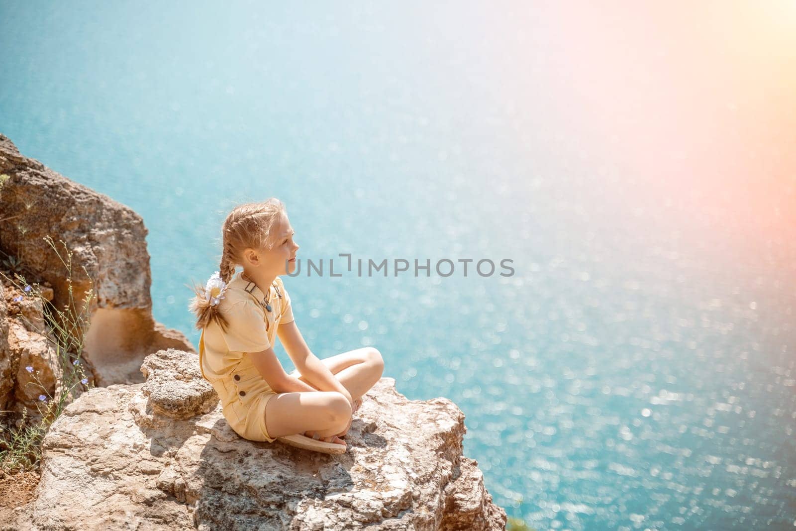 Happy girl perched atop a high rock above the sea, wearing a yellow jumpsuit and braided hair, signifying the concept of summer vacation at the beach