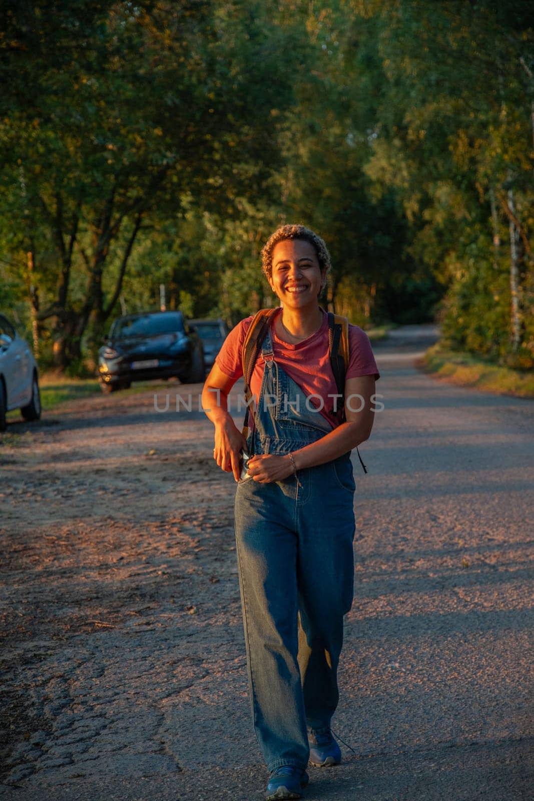 A young Brazilian woman in denim overalls walks along a forest road, the girl smiles happily, a car in the backgraund by KaterinaDalemans