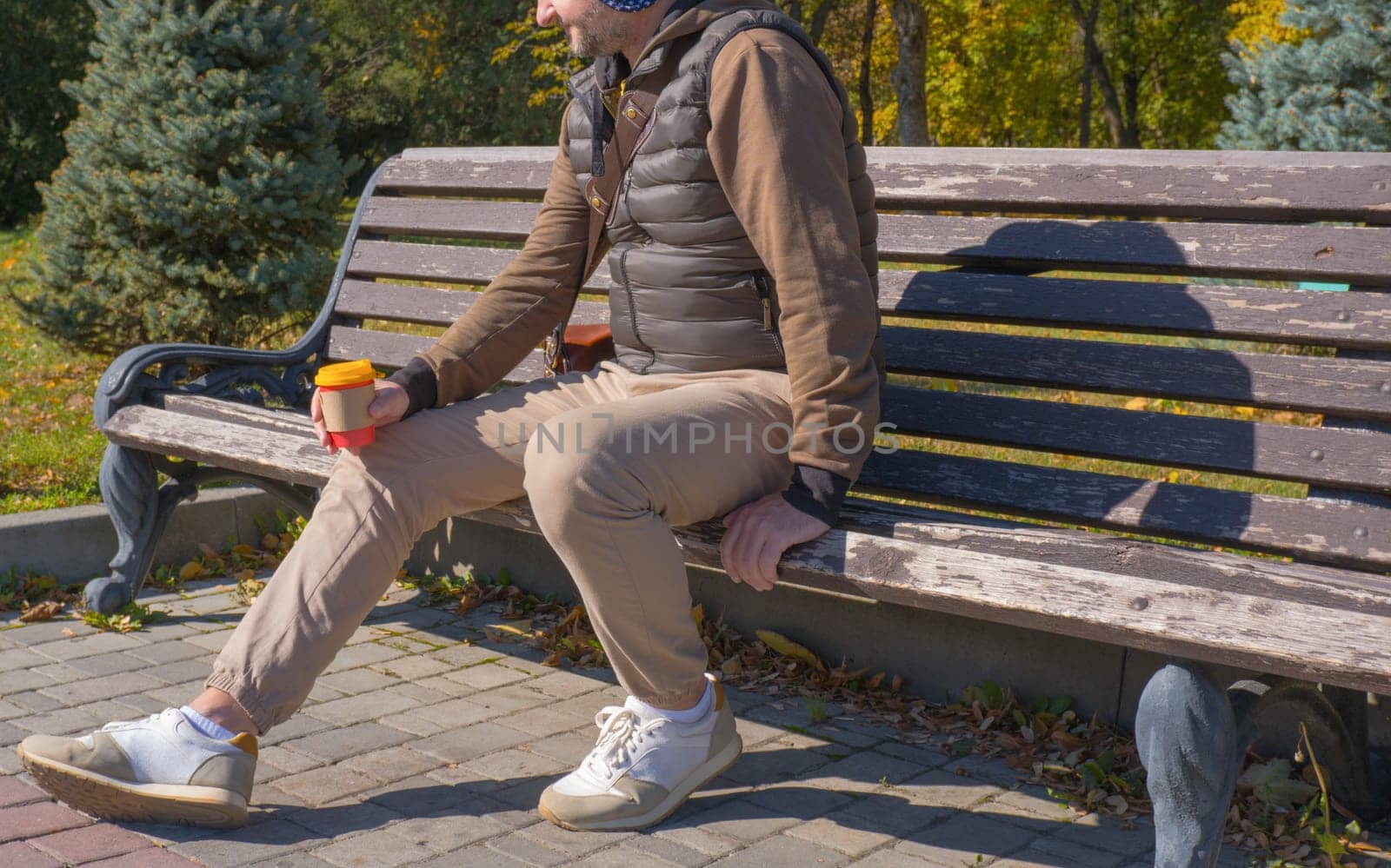 A young man sits on a bench in an autumn park, drinks coffee, rests, relaxes. Urban lifestyle concept. by Ekaterina34