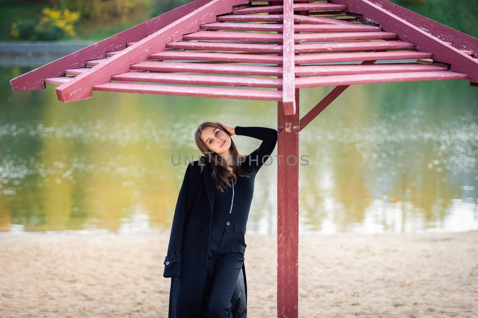 A beautiful girl poses while standing by a pond under an umbrella in an autumn park