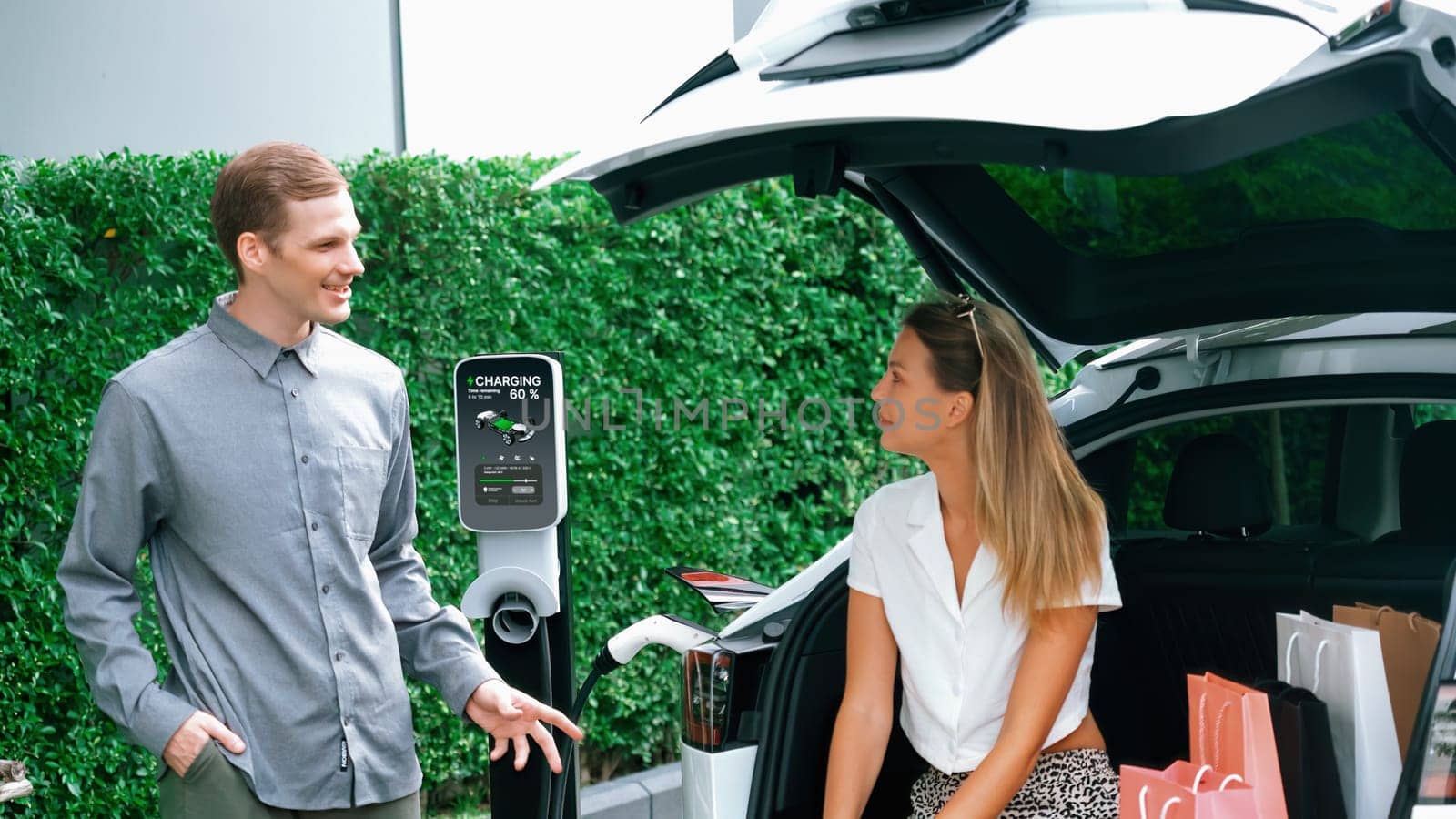 Young couple travel with EV electric car charging in green sustainable city outdoor garden in summer shows urban sustainability lifestyle by green clean rechargeable energy of electric vehicle innards