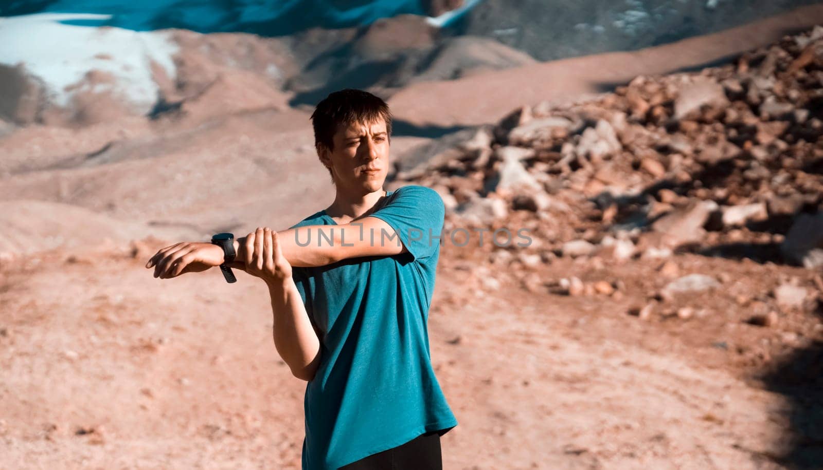 A young man does a warm-up before a trail and canyon race in the snow-capped mountains. The runner trains outdoor, does exercises in beautiful area.