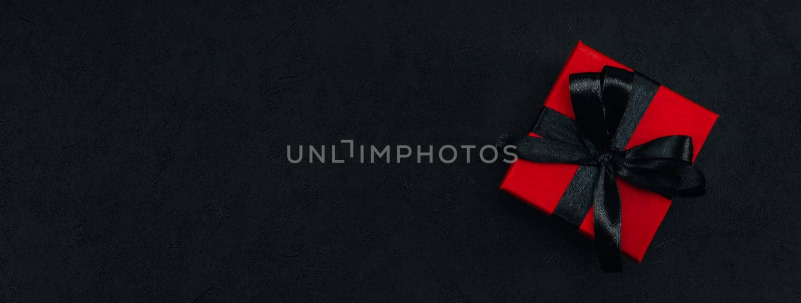 One red small gift box with a black ribbon tied in a bow lies on the right against a black background with space for text on the left, close-up top view. Christmas and black friday concept.