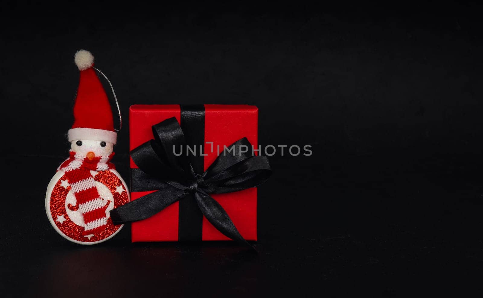 One red gift box and decorative snowman lies on the left against a black background with space for text on the right, close-up side view. Concept black friday, christmas.