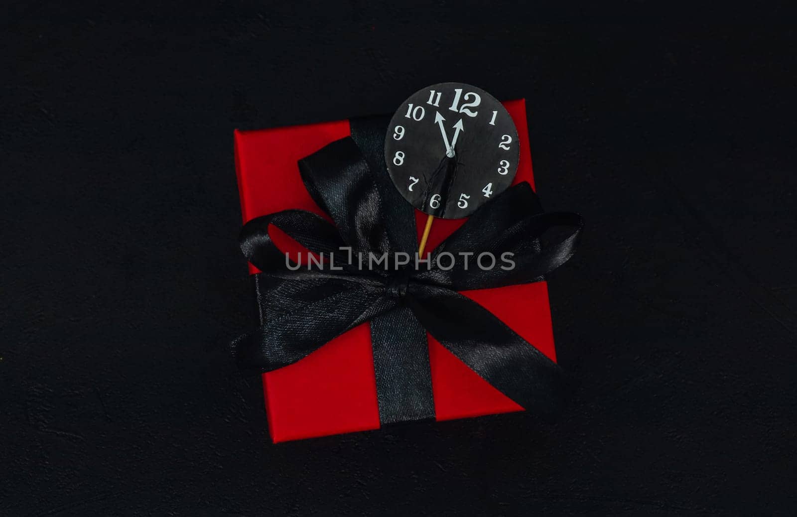 One small red gift box with a black ribbon and a paper clock in the middle on a black background with space for text, close-up top view. Christmas and black friday concept.