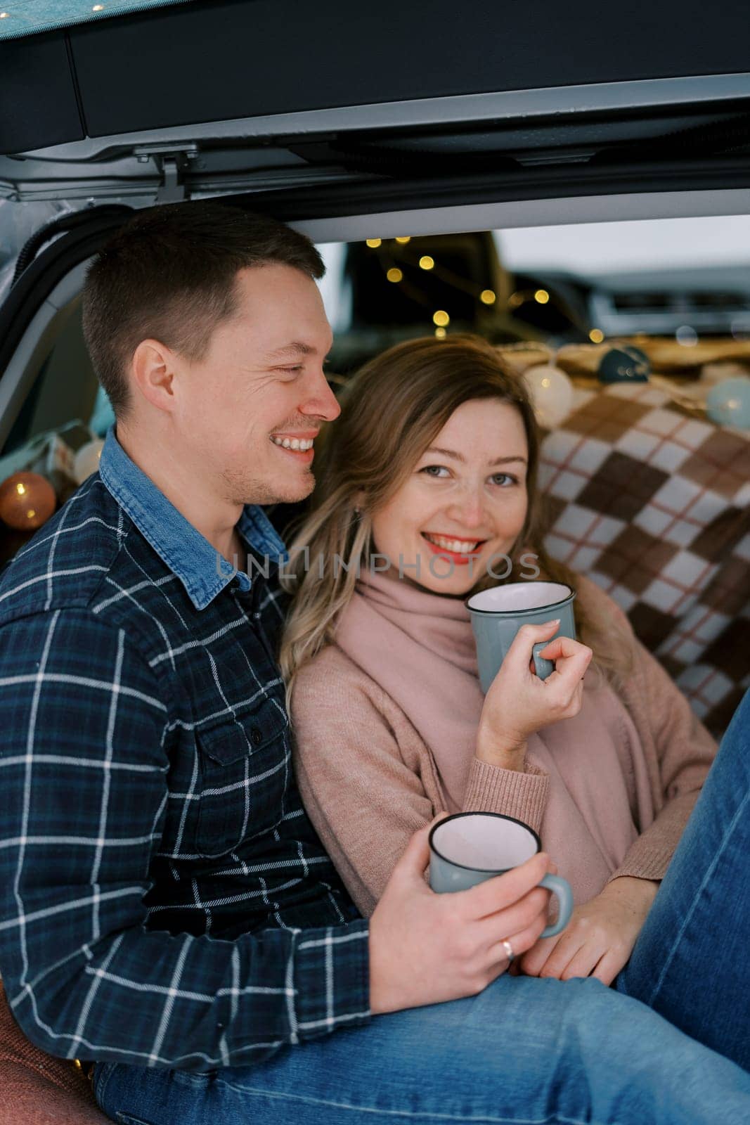 Laughing husband and wife with mugs of tea in their hands sit leaning against each other in the trunk of a car. High quality photo