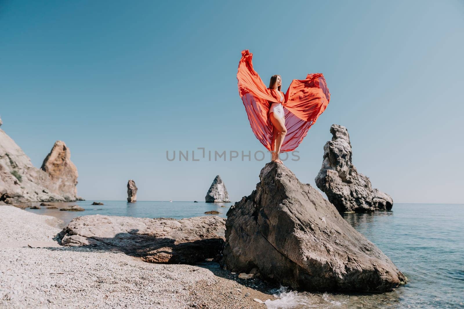 Woman travel sea. Young Happy woman in a long red dress posing on a beach near the sea on background of volcanic rocks, like in Iceland, sharing travel adventure journey by panophotograph