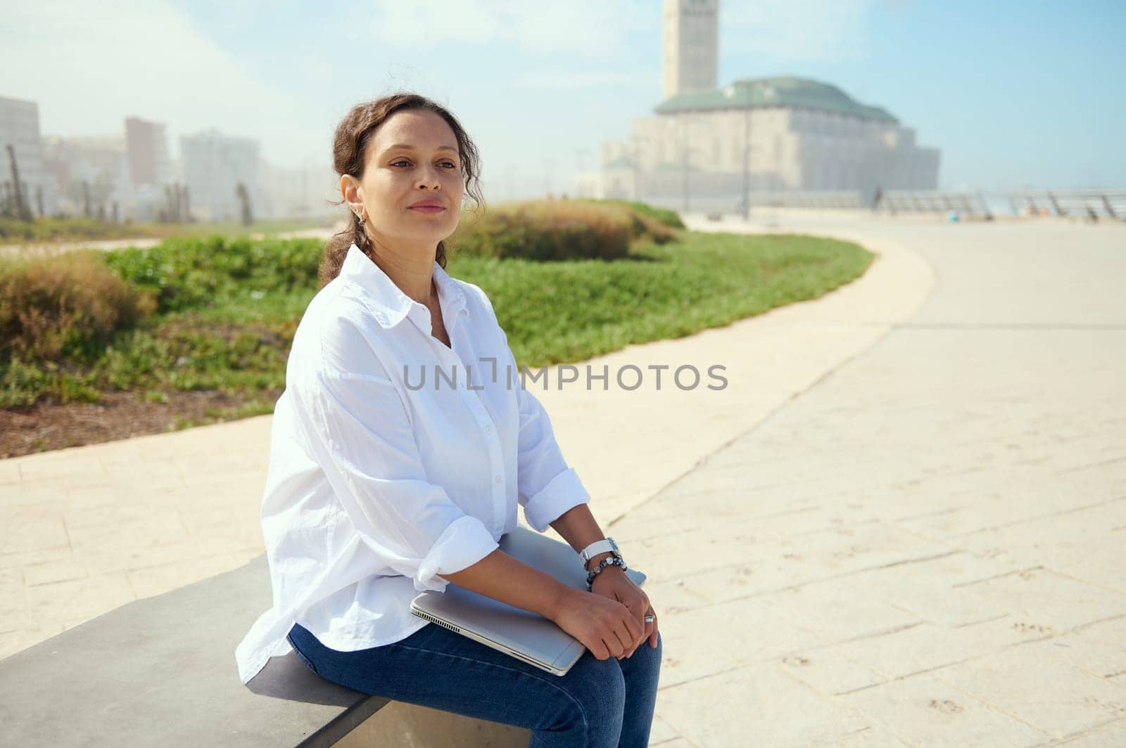 Confident business woman, freelance entrepreneur sitting on bench outdoors against a Hassan II mosque background, thinking on new startup project, dreamily looking into the distance. People. Lifestyle
