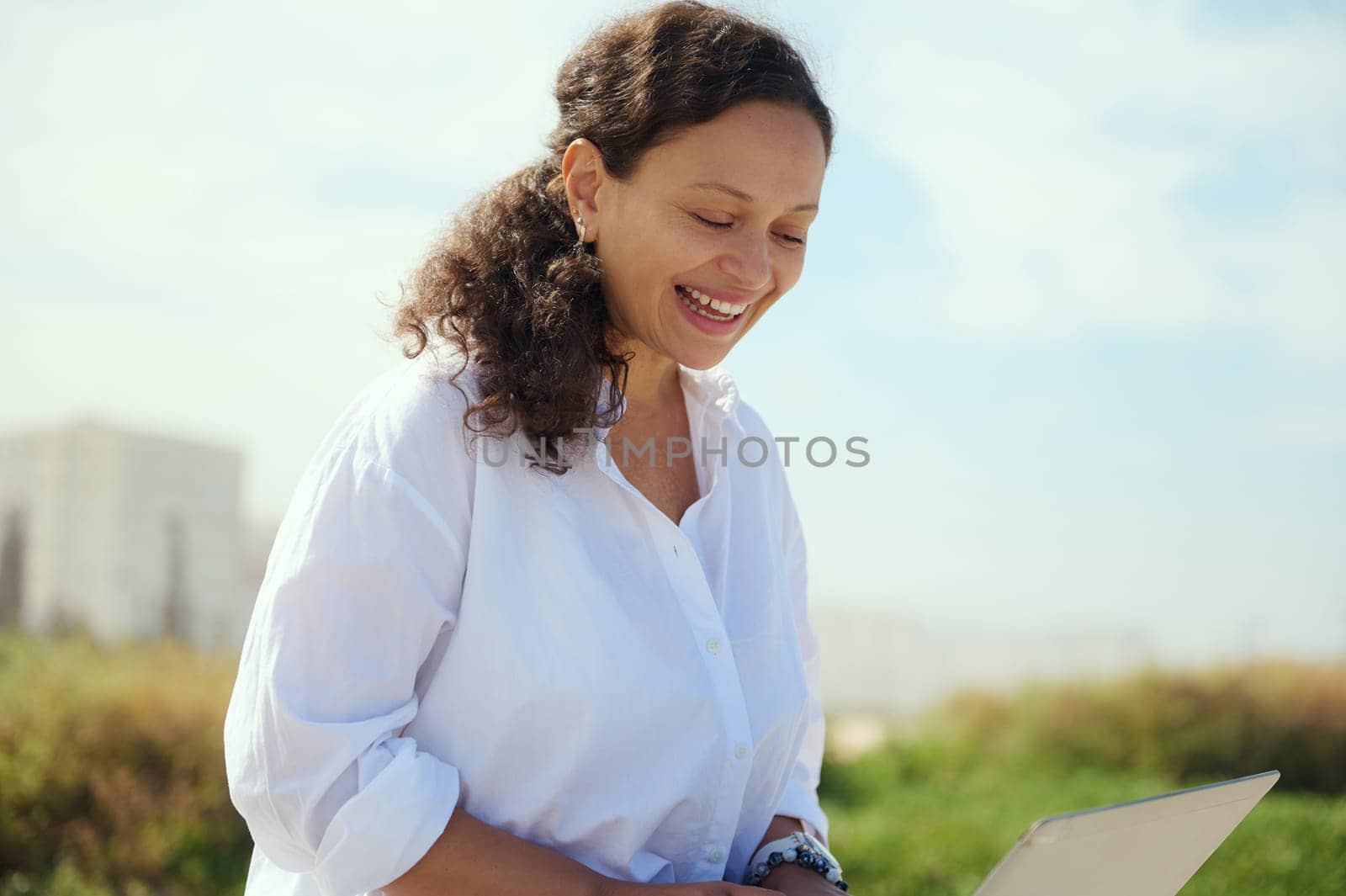 Beautiful multi ethnic young female manager smiling broadly while online working outdoors, using laptop. Confident closeup portrait of a businesswoman expressing positive emotions, enjoying remote job