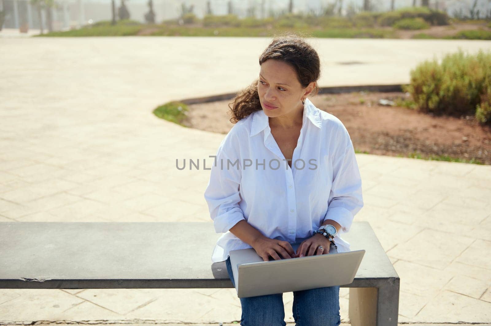 Confident pensive business woman thinking on new startup project, online working on laptop, sitting on bench outdoors by artgf