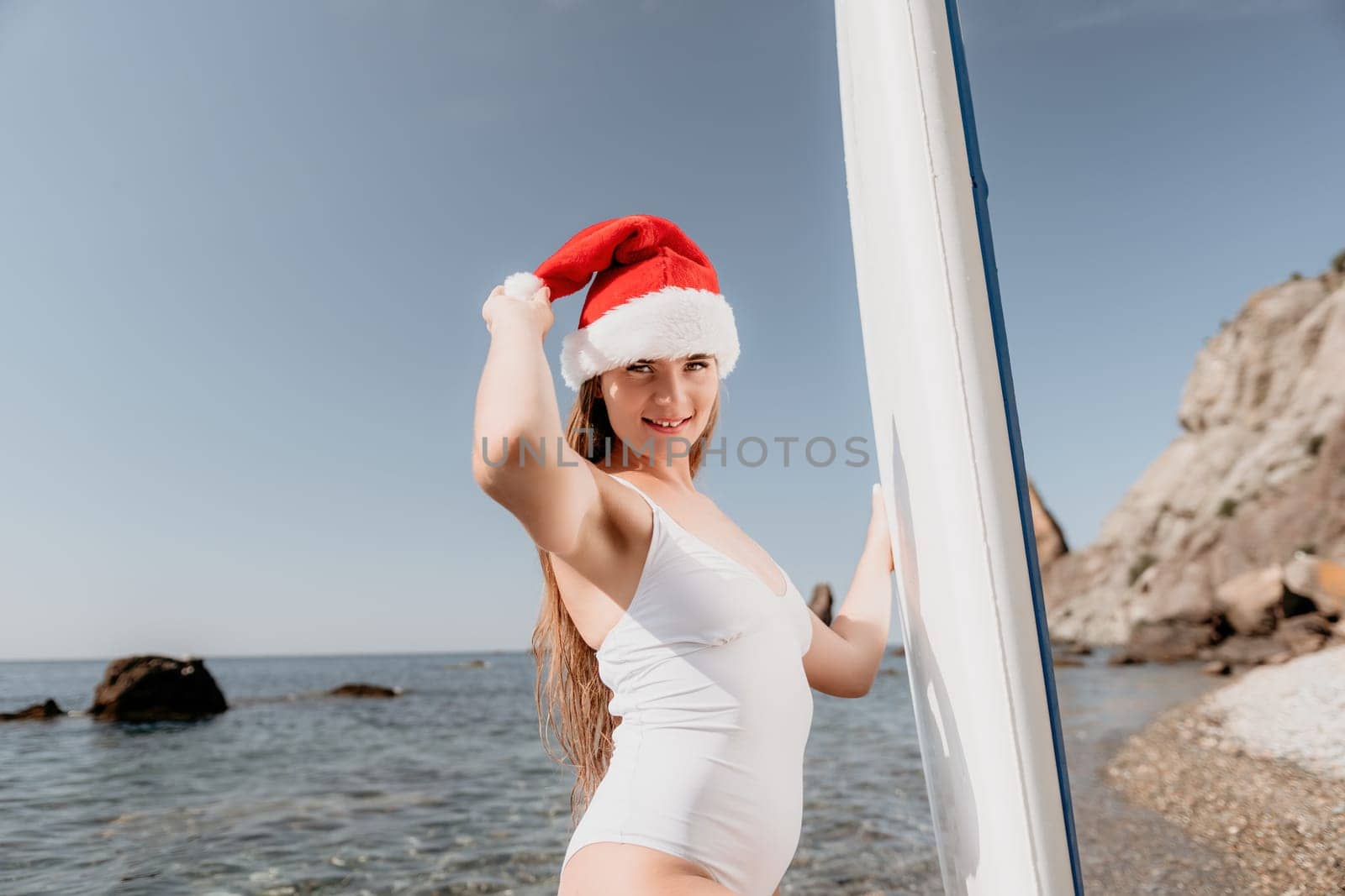 Woman sea sup. Close up portrait of happy young caucasian woman with long hair in Santa hat looking at camera and smiling. Cute woman portrait in a white bikini posing on sup board in the sea by panophotograph