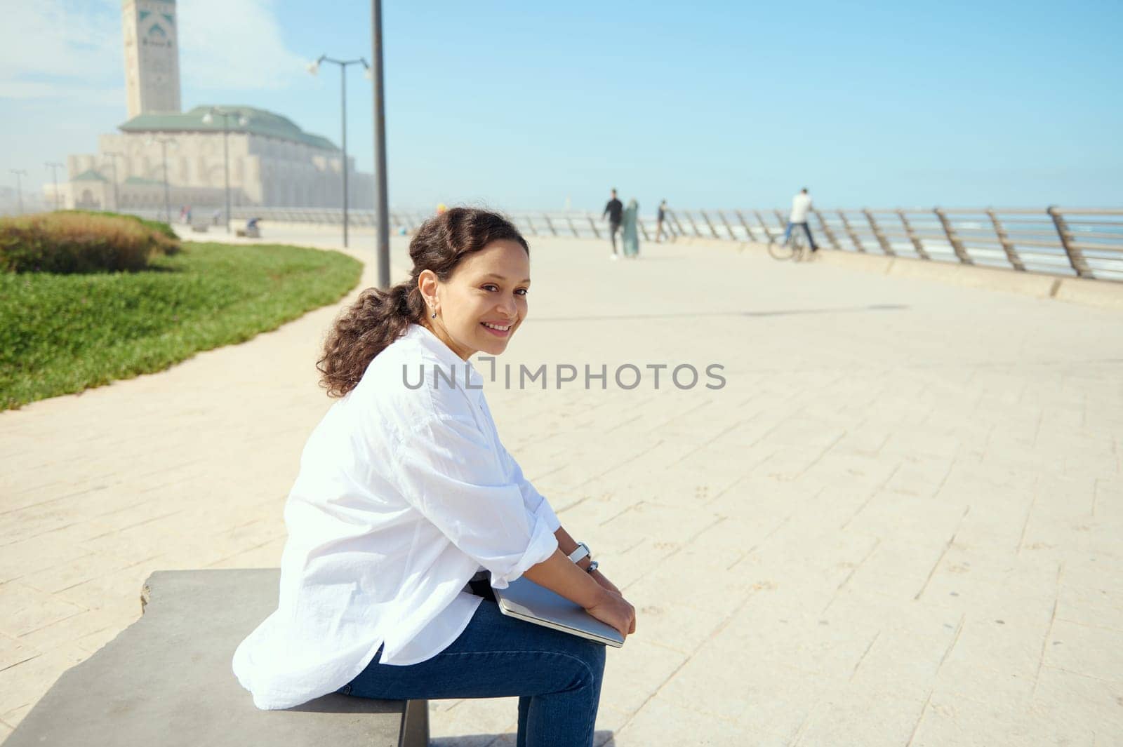 Confident portrait of a charming young adult woman in casual clothes, sitting on a bench outdoors, holding laptop and cutely smiling with a beautiful toothy smile, looking dreamily into the distance