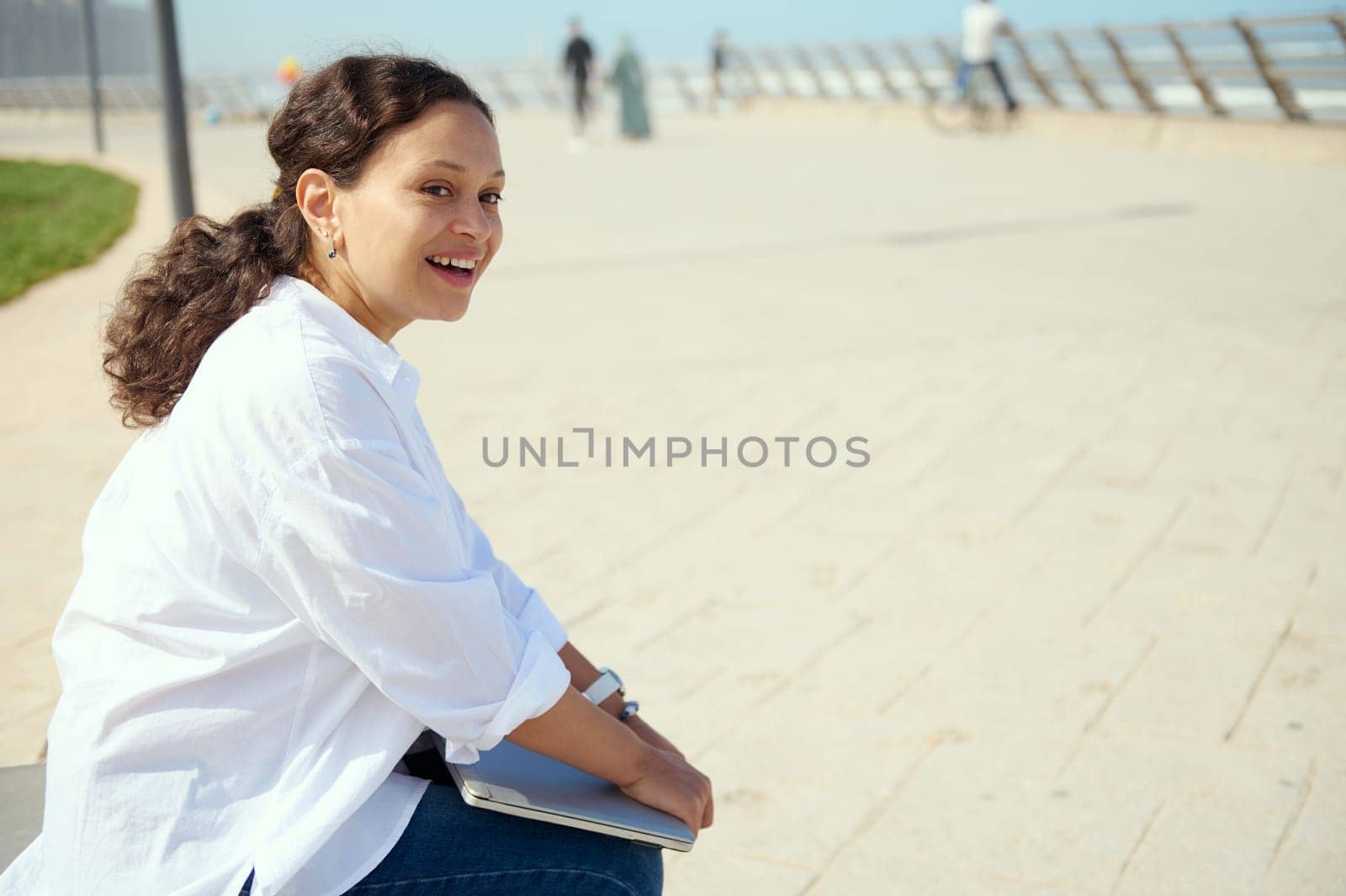 Multi ethnic young freelance entrepreneur woman holding laptop while resting outdoors after work, smiling broadly looking aside. Charming purposeful businesswoman telecommuting outdoors.