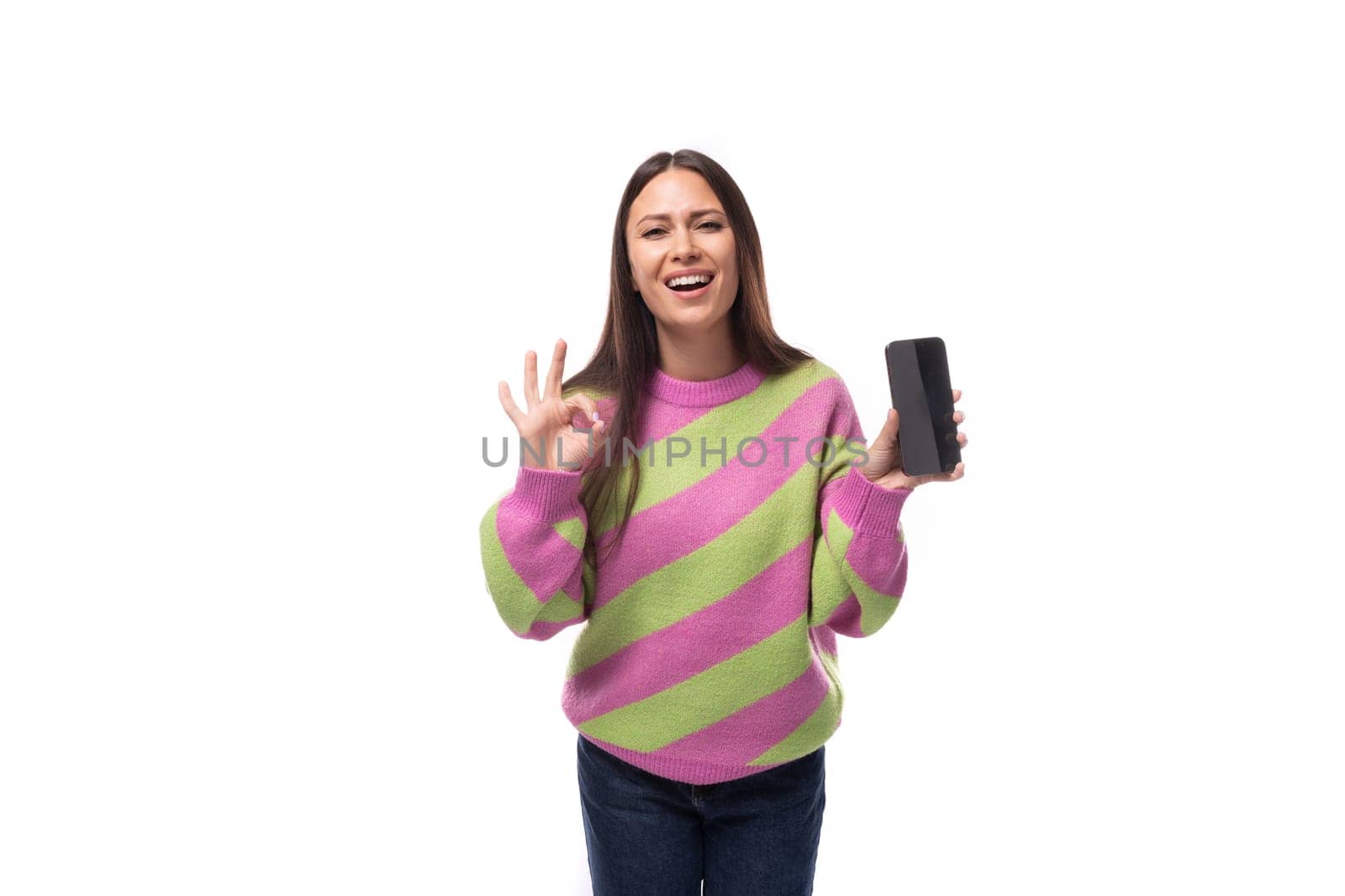 beautiful young brunette woman in a striped pink blouse shows the screen of a smartphone with a mockup on a white background with copy space.
