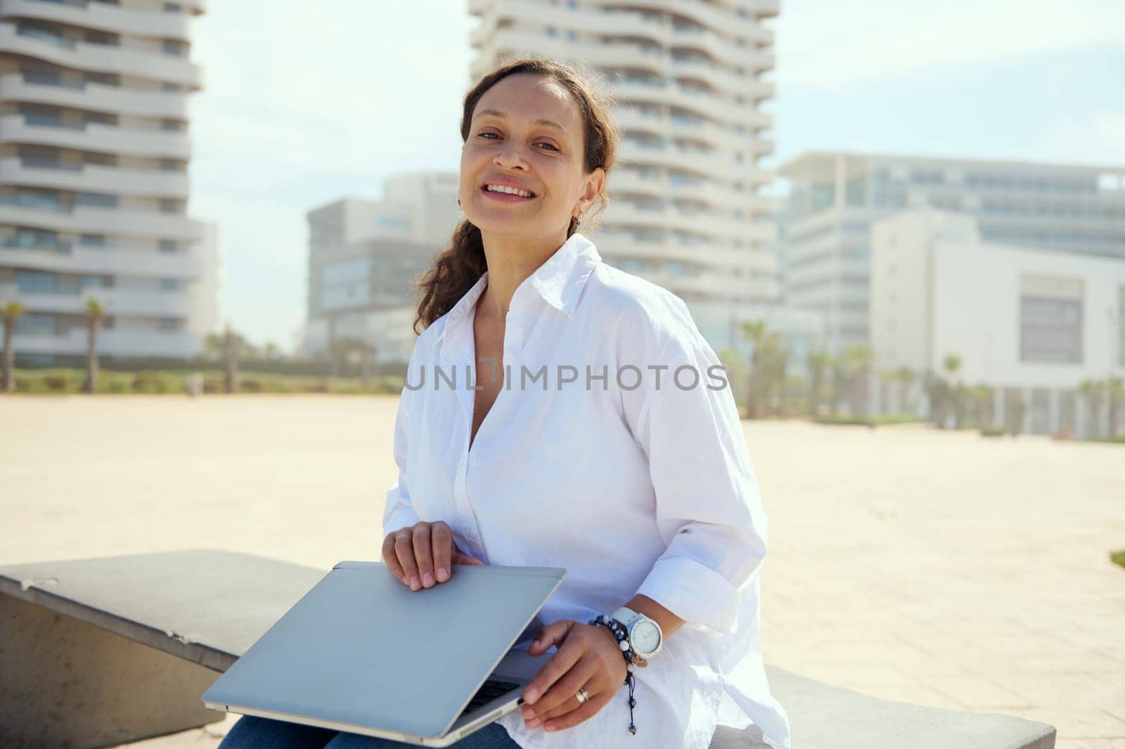 Authentic multi ethnic business lady, freelance entrepreneur, holding laptop and smiling looking at camera, sitting on bench outdoors. People. Career. Recruitment. Online business. Remote job