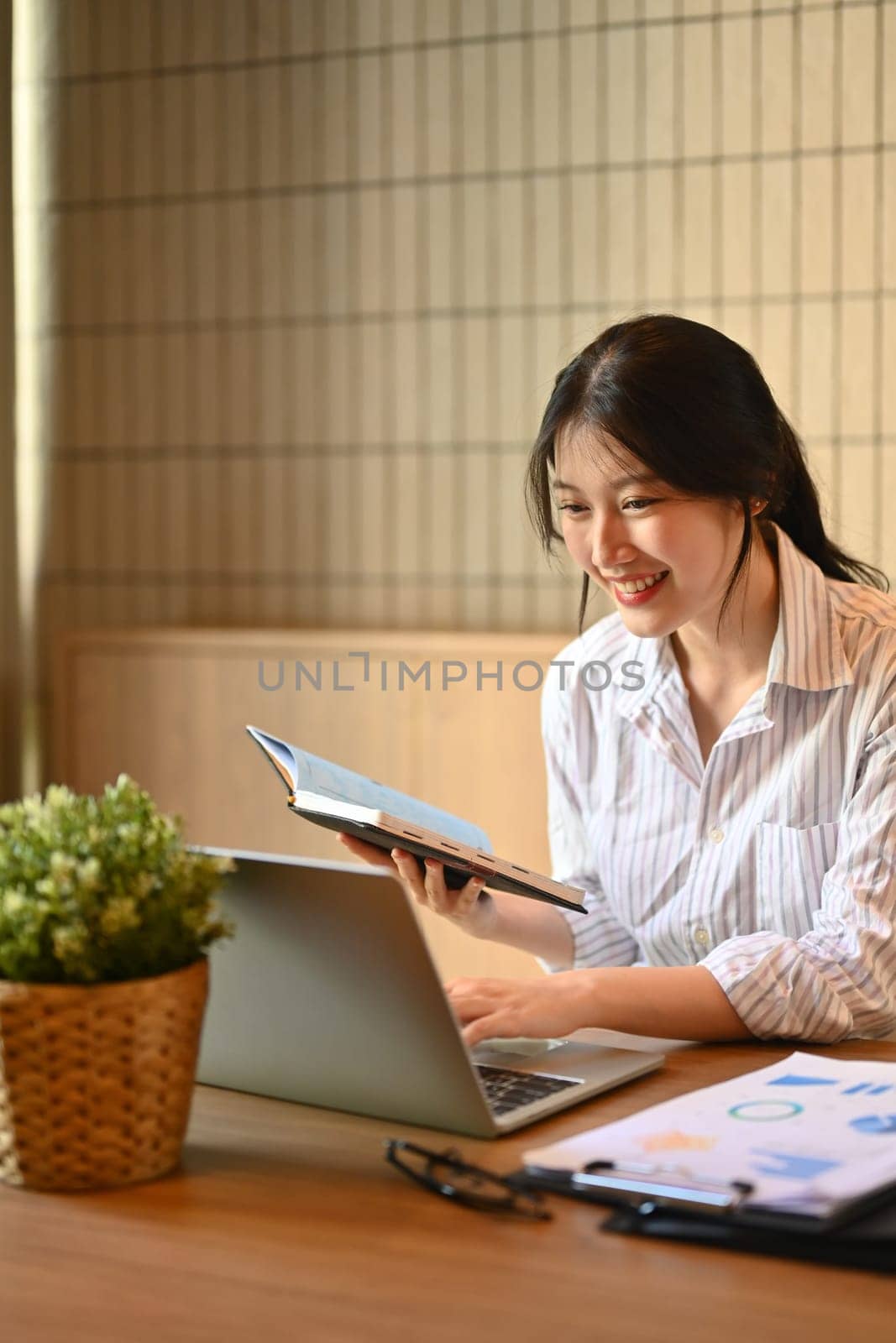 Beautiful young businesswoman holding notepad and checking information on laptop computer.