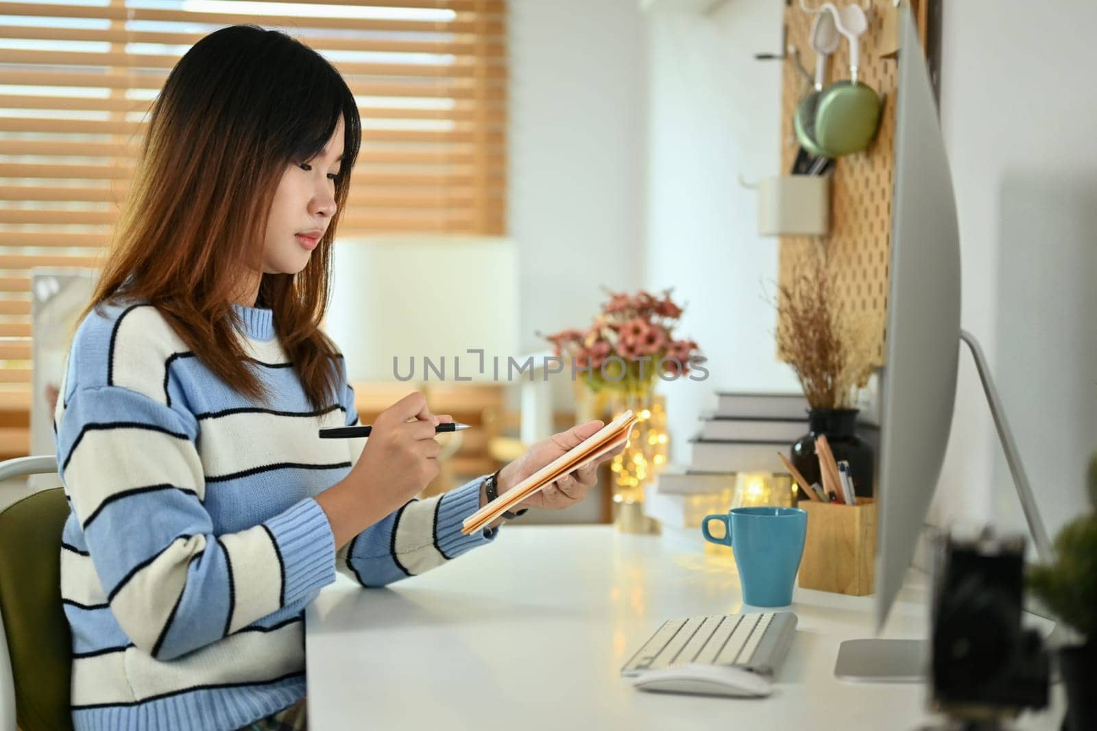 Young woman sitting in front of computer monitor and writing notes on notepad.