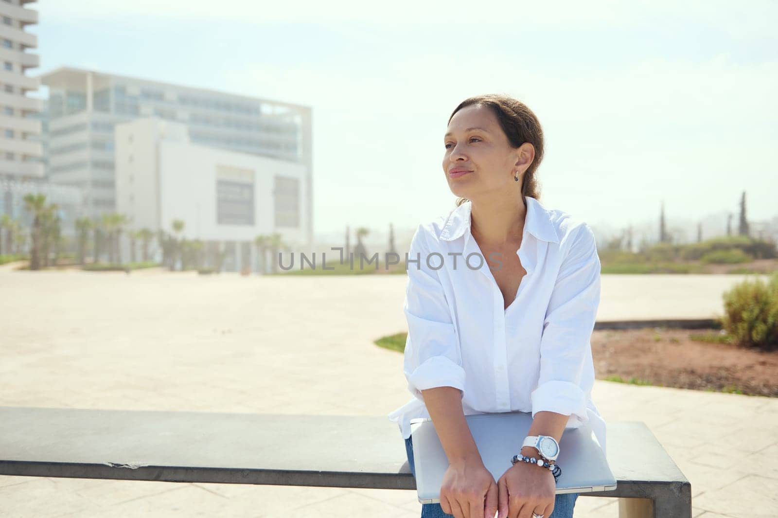 Authentic confident portrait of young adult multi ethnic woman, business lady, freelancer, entrepreneur, developer sitting on a bench against cityscape background and looking aside dreamily