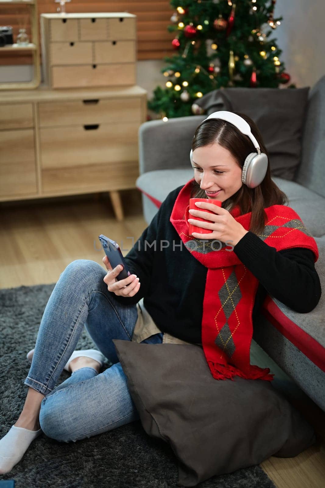 Portrait of caucasian woman reading message on mobile during winter sitting on floor against Christmas tree with lights.