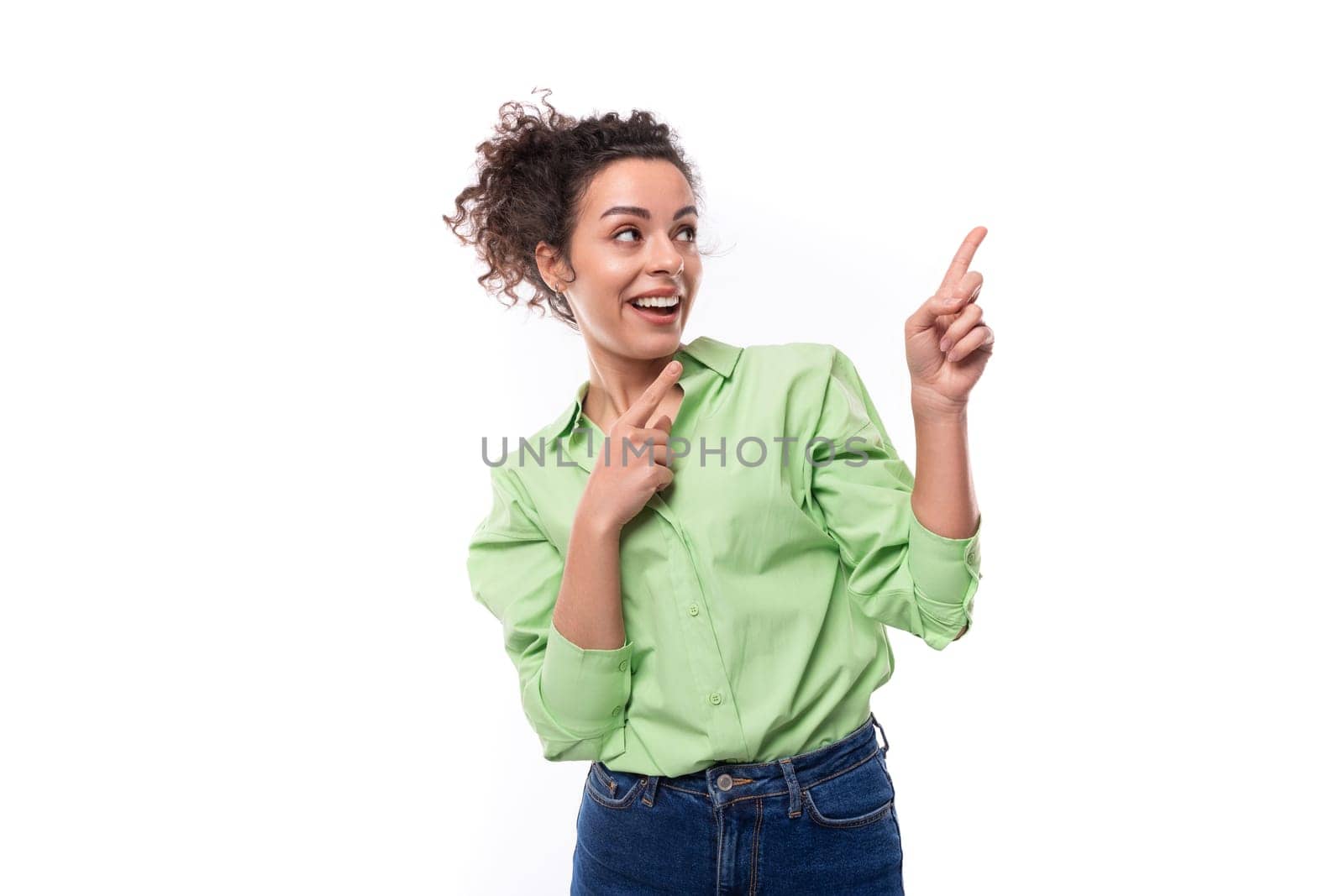 young brunette curly slender female model dressed in a light green shirt points with her hand at the space of the wall.