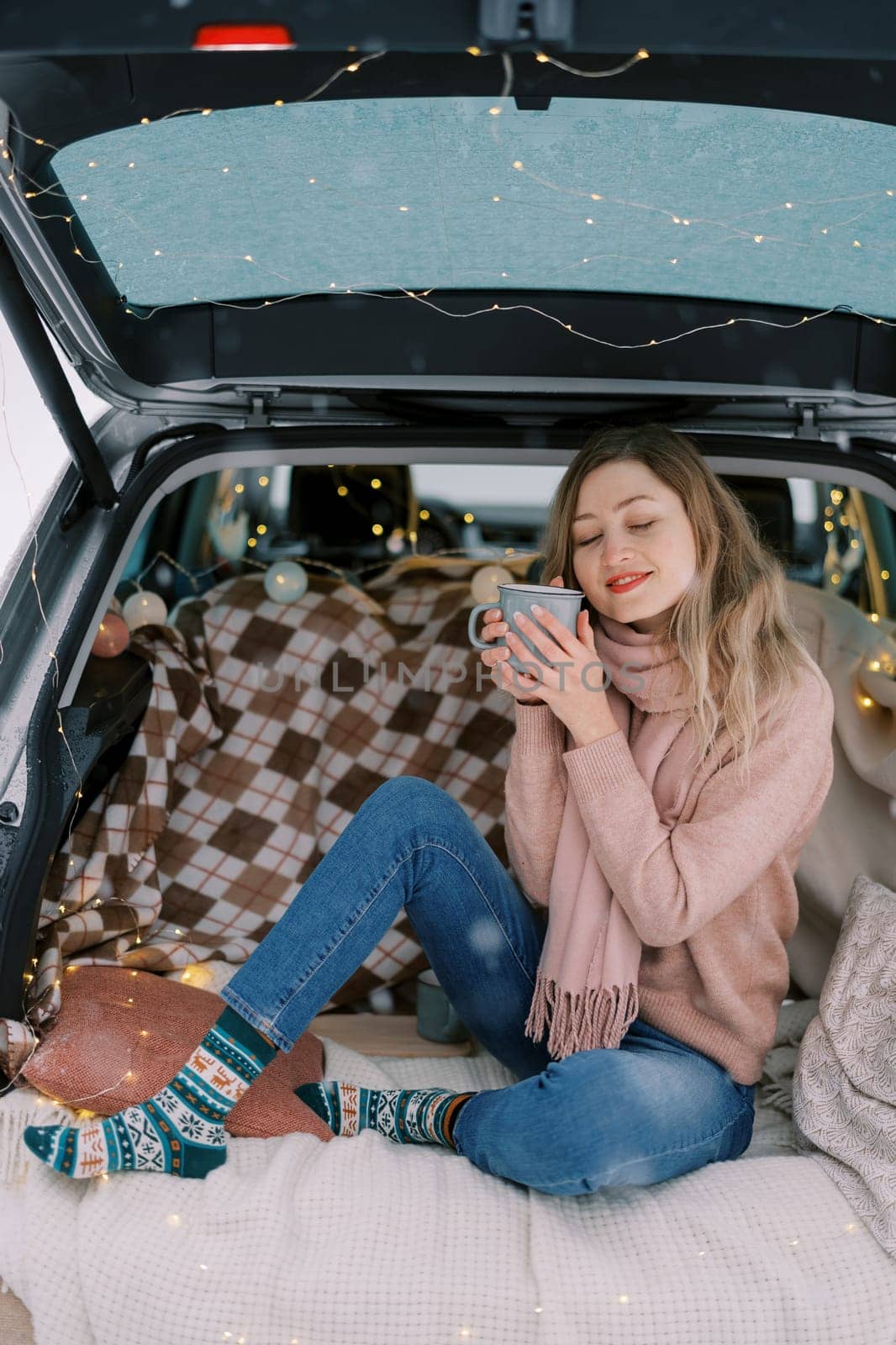 Smiling girl sitting with mug of coffee squinting in car trunk in winter forest. High quality photo