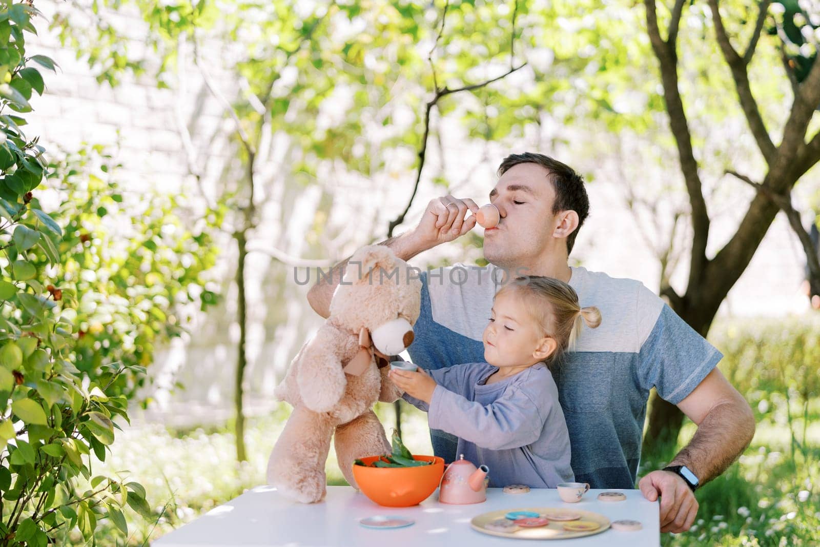 Little girl treats a teddy bear to tea from a toy cup while sitting next to her dad drinking from a toy cup. High quality photo