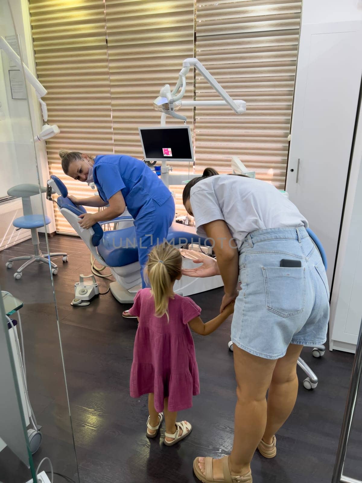 Tivat, Montenegro - 06 august 2023: Mom with a little girl stand near the dentist adjusting the dental chair by Nadtochiy