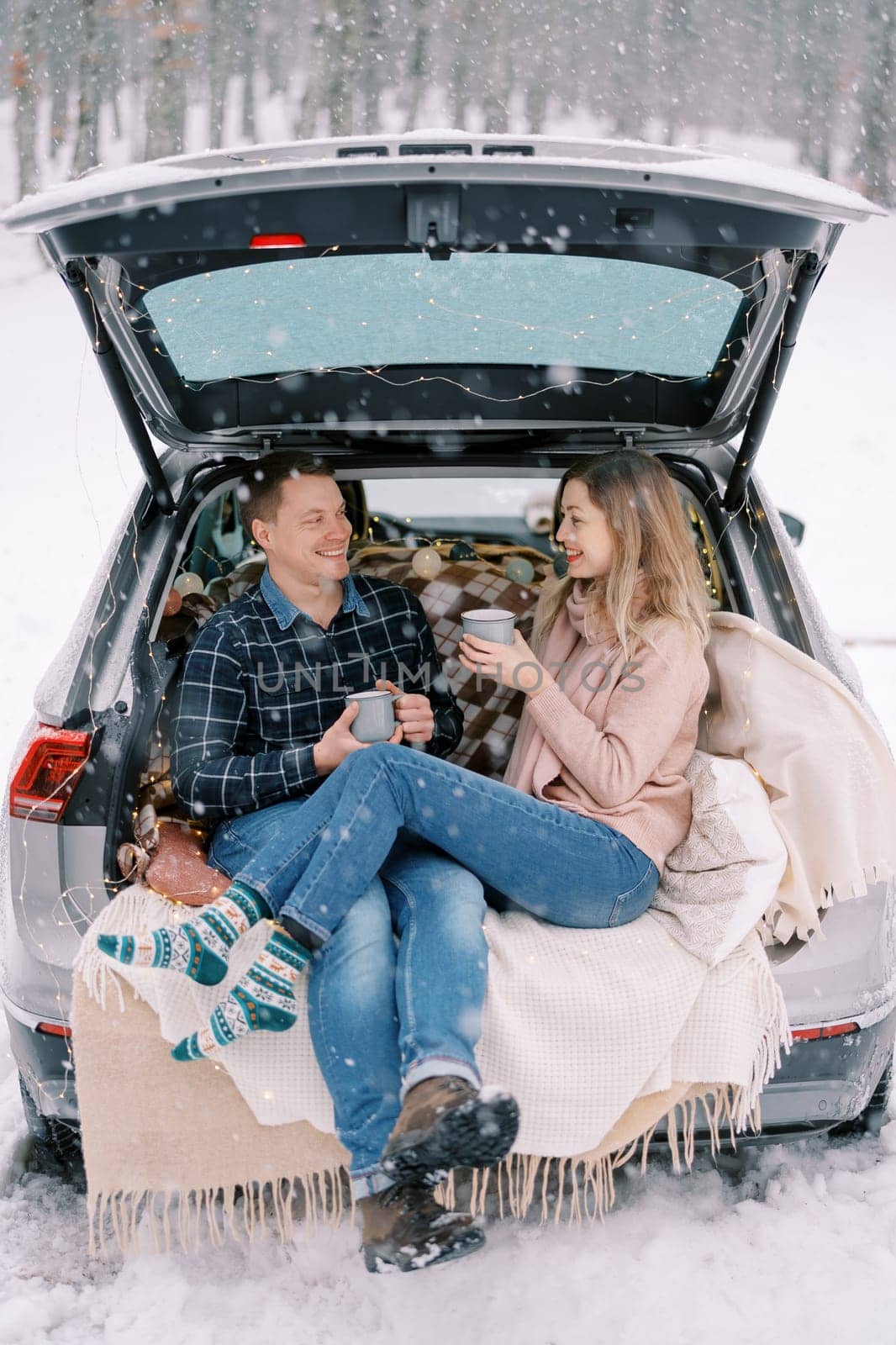 Smiling man and woman sitting with cups in car trunk on blankets in snowy park. High quality photo