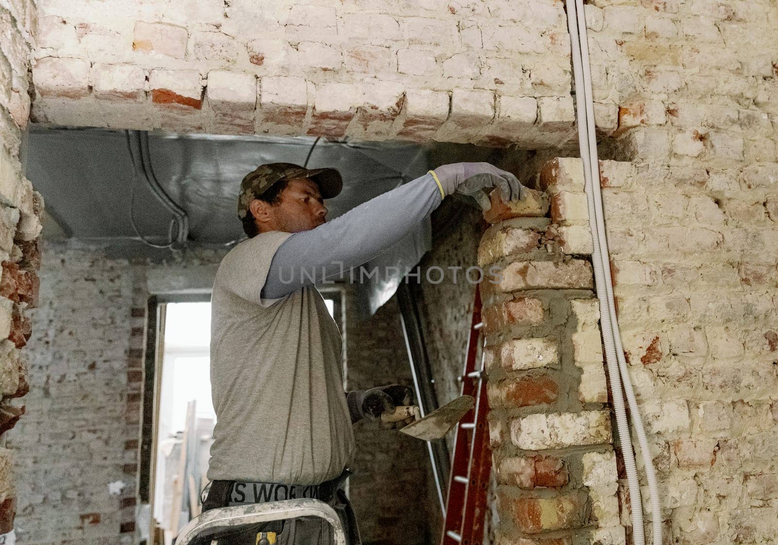 One young Caucasian man laying bricks on fresh cement in a doorway, standing on a stepladder to the right, close-up side view.