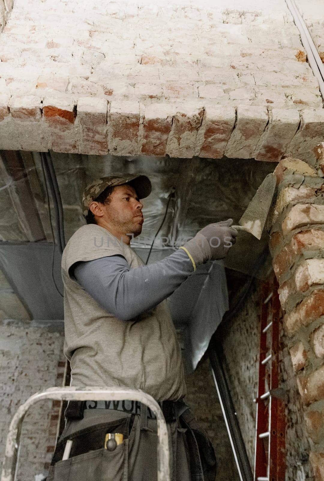 One young Caucasian man spreads fresh cement onto bricks using a spatula, leveling it and standing during the day on the right on a stepladder in an old house, close-up view from below.