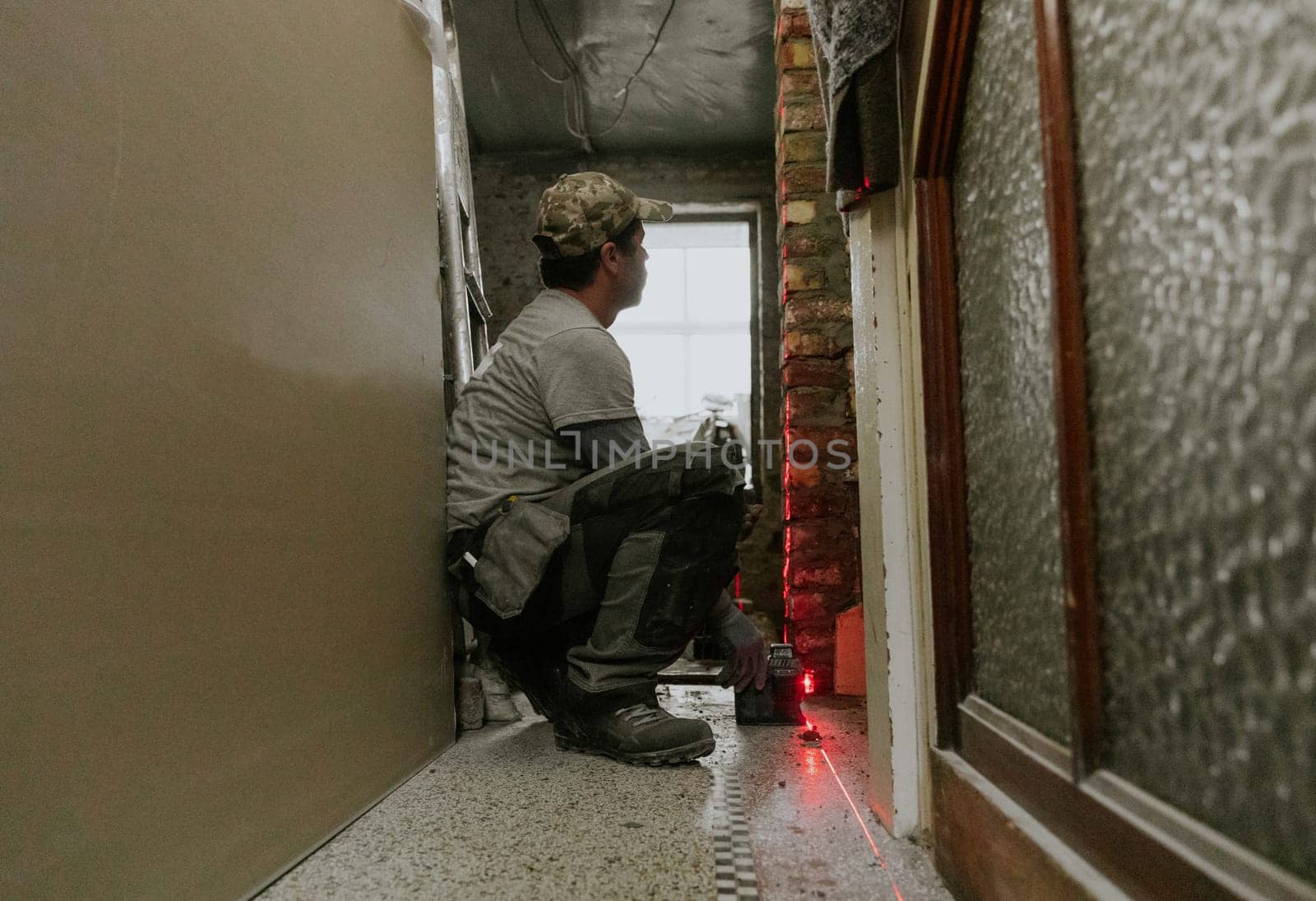 One young Caucasian man in casual clothes sits on his haunches, looks down the street and measures fresh brickwork in a doorway in the corridor of an old house using a laser level, close-up view from below.