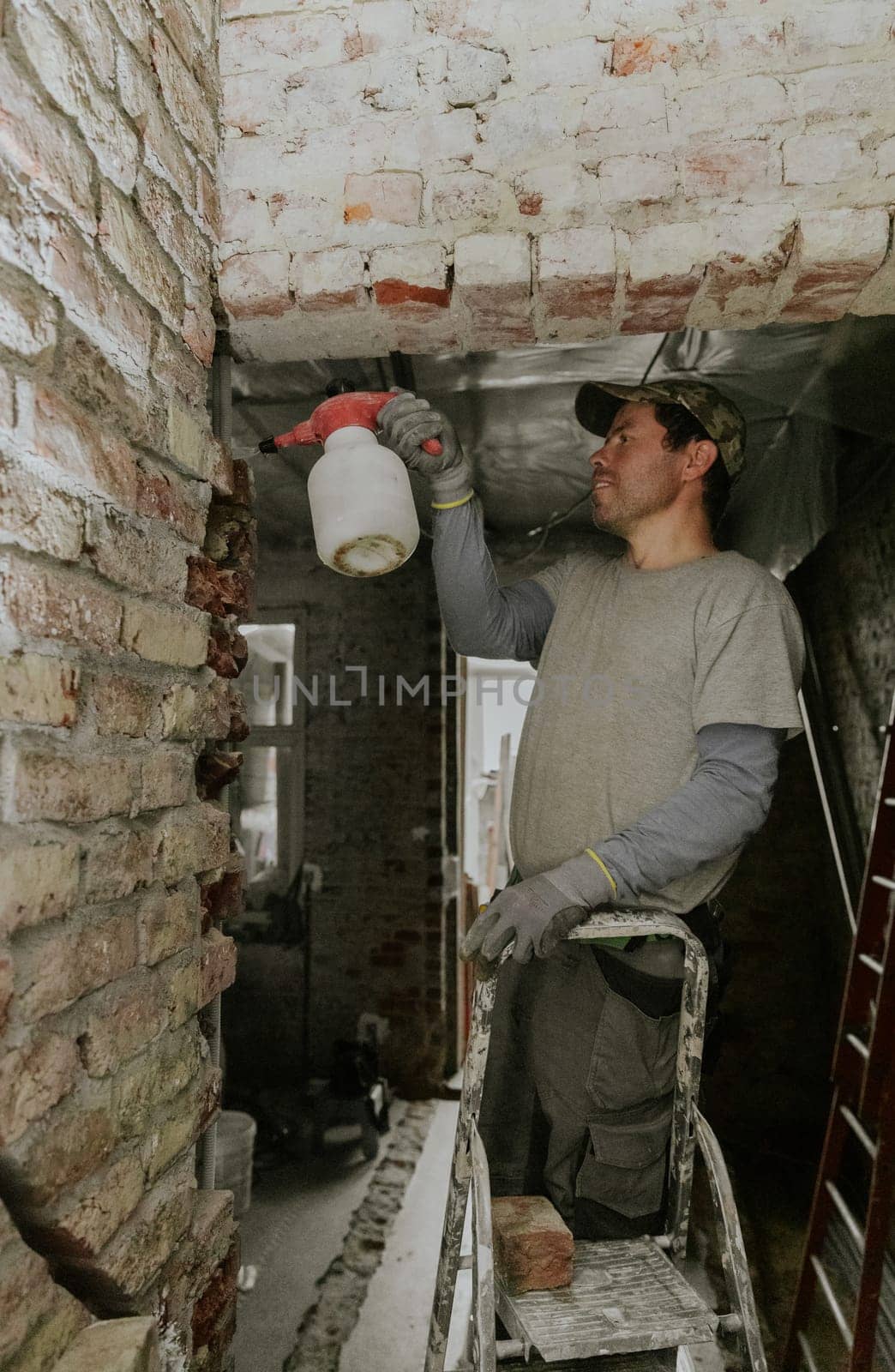 One young handsome Caucasian happy brunette man in casual clothes stands on a stepladder and sprays water from a spray bottle onto bricks in a doorway under the ceiling in an old house, close-up view from below.