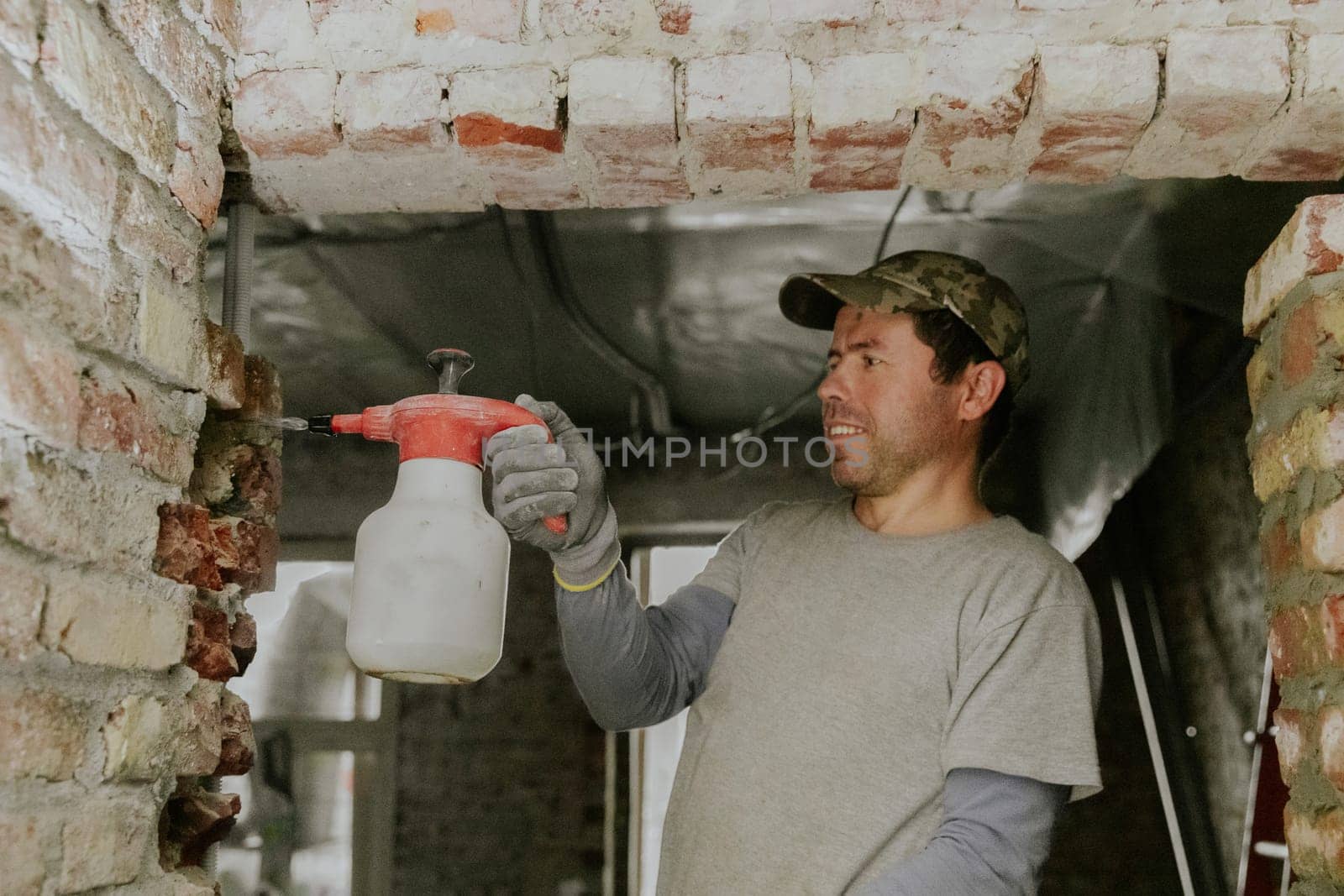 One young handsome Caucasian happy brunette man in casual clothes stands on a stepladder and sprays water from a spray bottle onto bricks in a doorway under the ceiling in an old house, close-up side view.