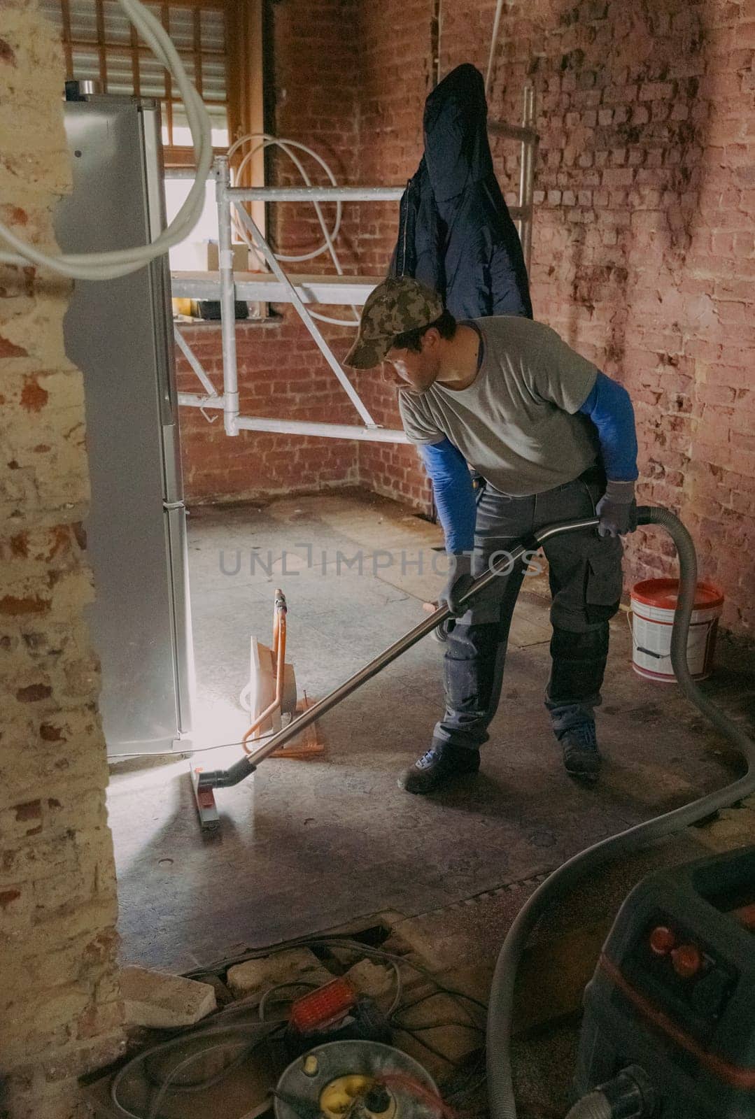 One young Caucasian man in a gray casual work uniform vacuums up construction debris after laying bricks in a room where renovations are underway, standing sideways under the light of a lantern in an old ruined house, close-up side view.