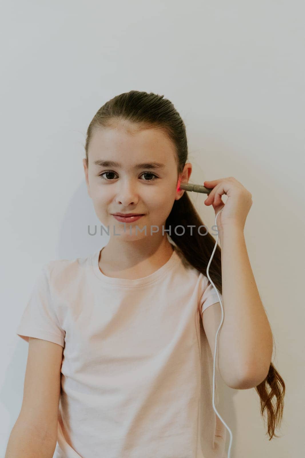 One beautiful Caucasian brunette girl with collected hair and in a pink T-shirt treats her ear with an infrared light device, standing against a white wall, close-up side view.