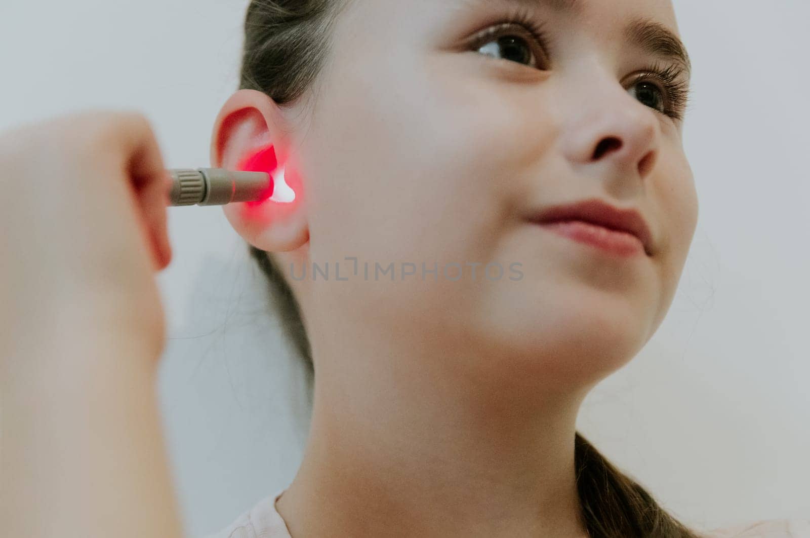 One beautiful Caucasian brunette girl with a happy smile, collected hair and in a pink T-shirt treats her right ear with an infrared light device, sitting on a bed at home near a white wall, a very close-up view from below.