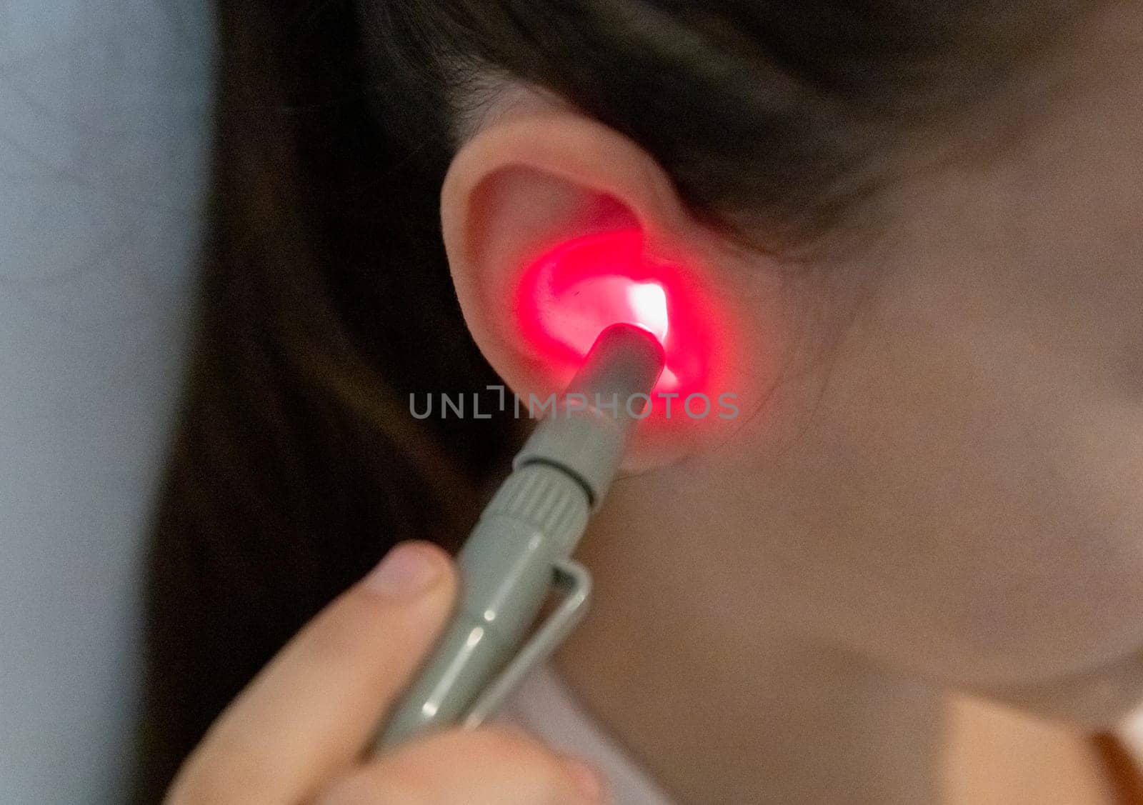 One beautiful Caucasian brunette girl with a happy smile, collected hair and in a pink T-shirt treats her right ear with an infrared light device, sitting on a bed at home near a white wall, top-side view very close-up.