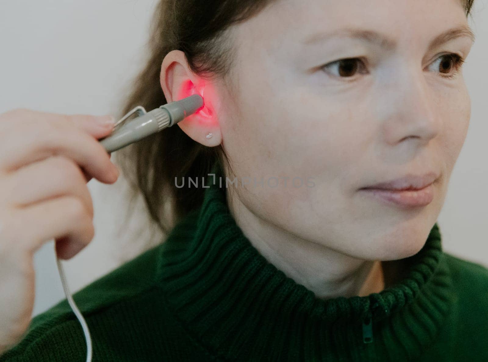 One beautiful Caucasian brunette girl with collected hair in a green sweater treats her right ear with an infrared light device, sitting on a bed against a white wall, side view very close-up.