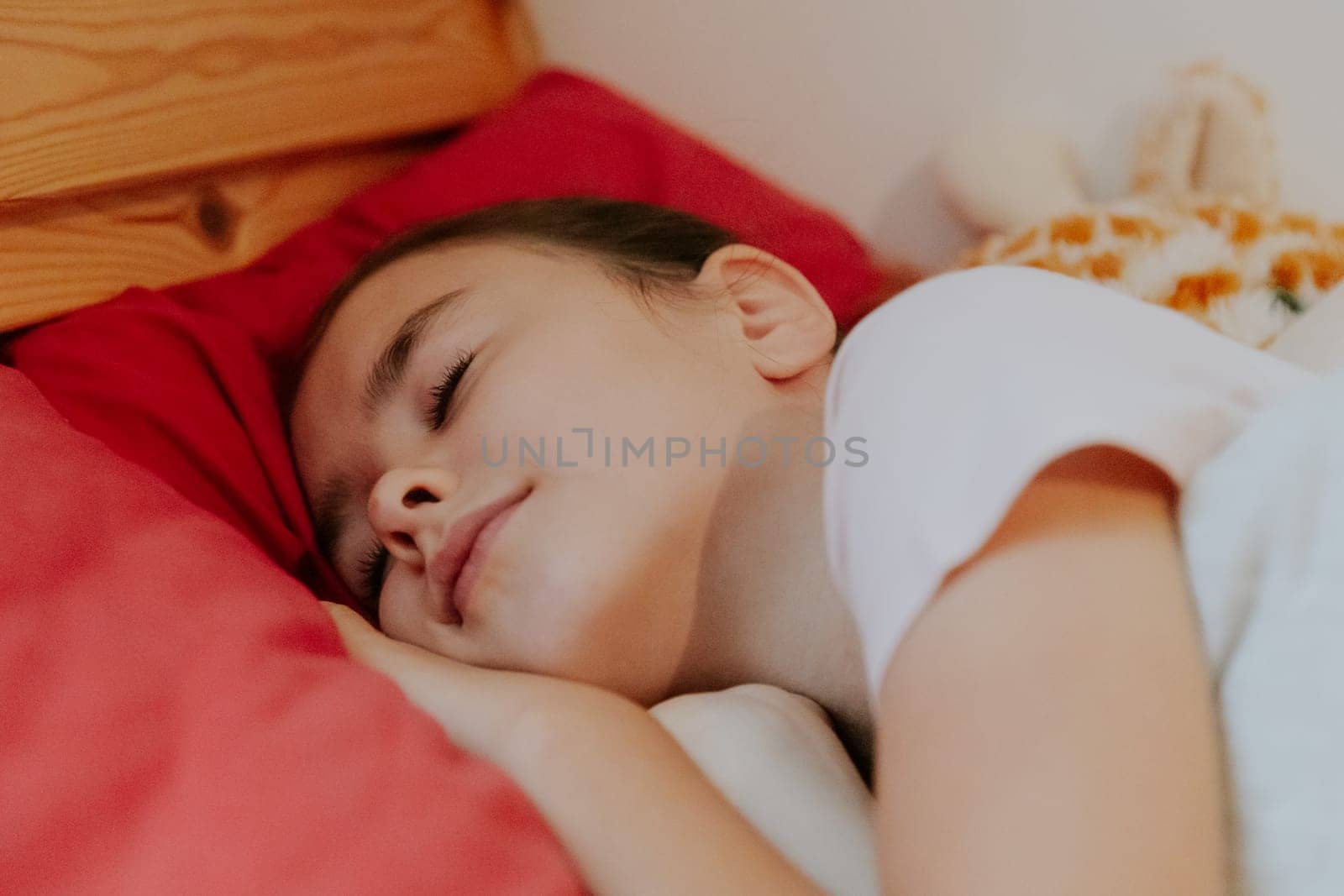 One beautiful little Caucasian girl sleeps sweetly in a wooden bed on a red pillow with a soft toy giraffe behind her back, close-up side view.