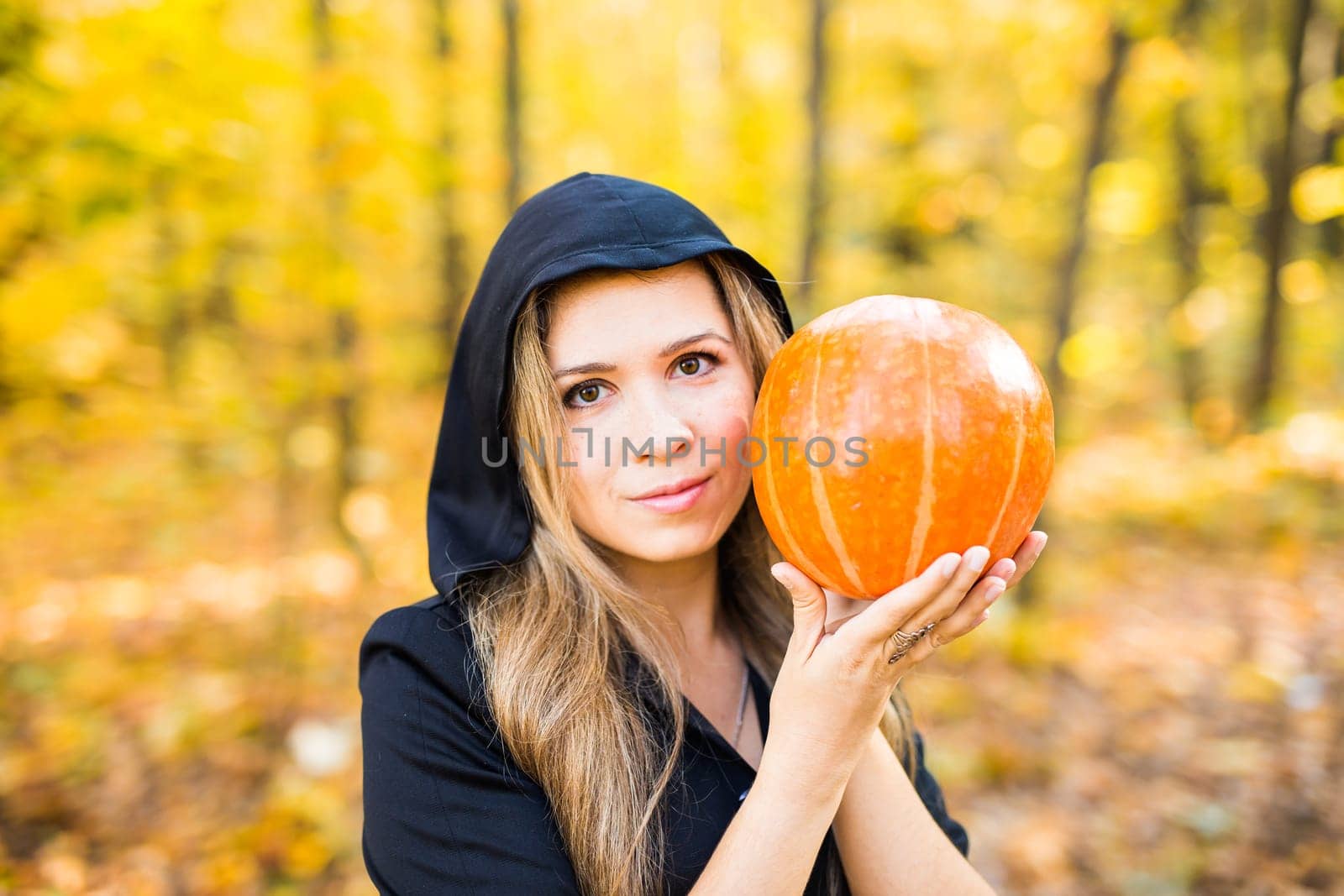 Halloween Witch with a magic Pumpkin in a dark forest. Beautiful young woman in witches hat and costume holding carved pumpkin. Halloween art design