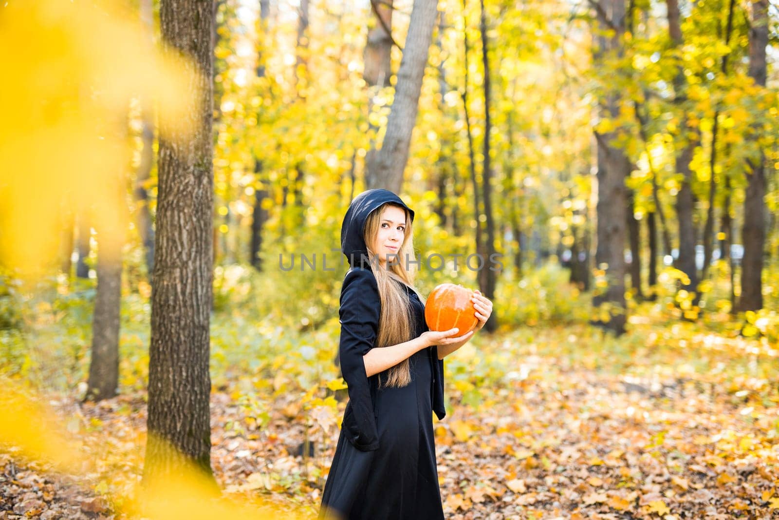 Halloween Witch with a magic Pumpkin in a dark forest. Beautiful young woman in witches hat and costume holding carved pumpkin. Halloween art design