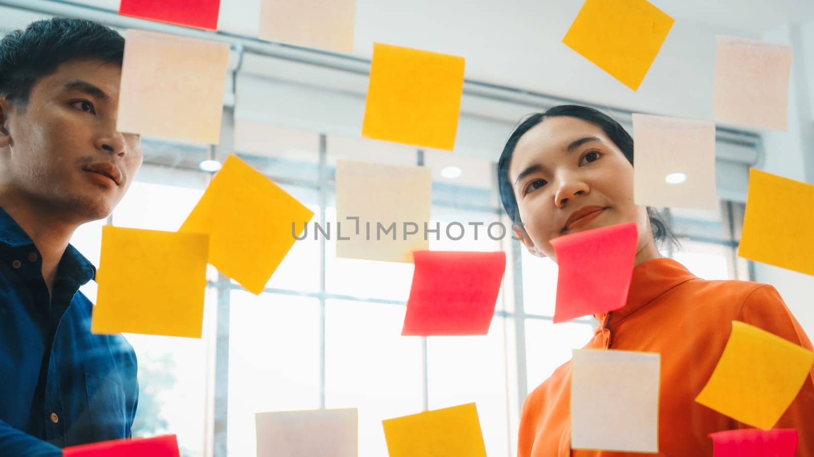 Business people work on project planning board in office and having conversation with coworker friend to analyze project development . They use sticky notes posted on glass to make it organized. Jivy