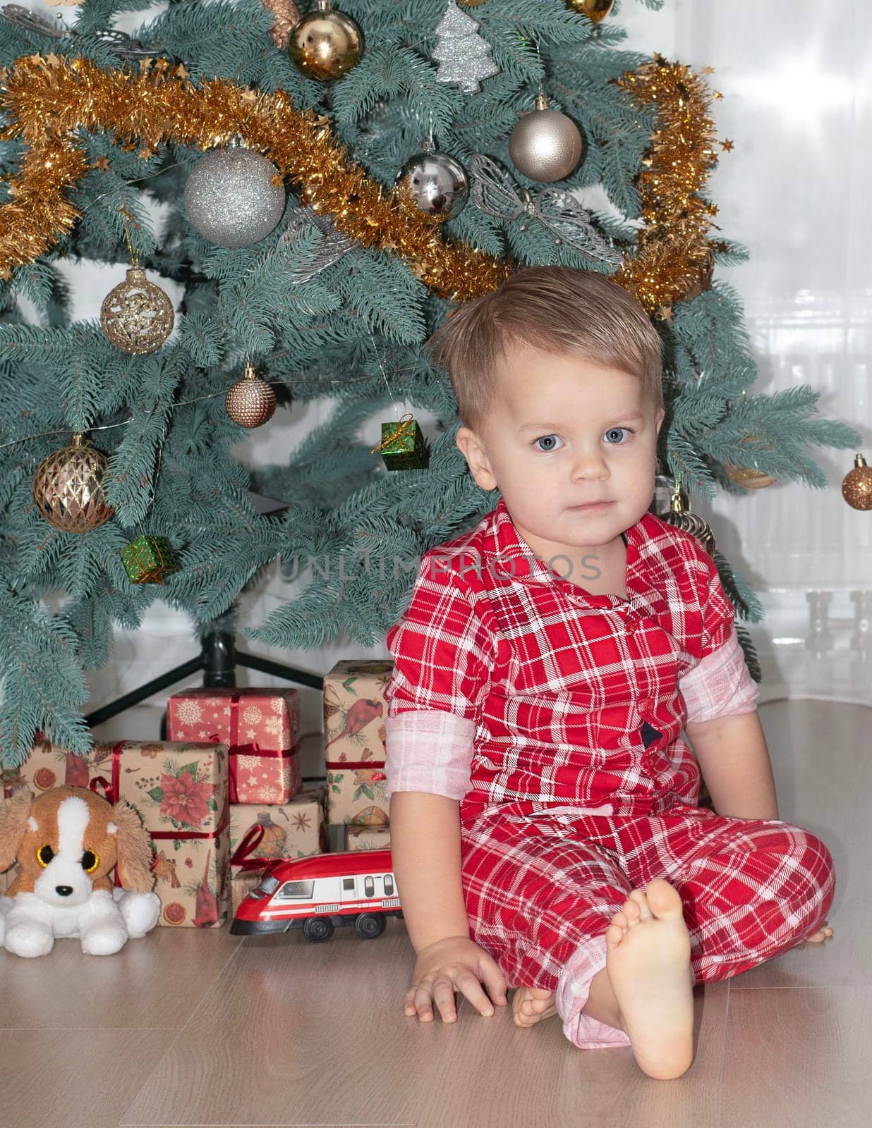 Christmas concept. New Year 2024. A small handsome boy in red checkered pajamas sits on the floor against the background of a decorated Christmas tree with gifts. Close-up.