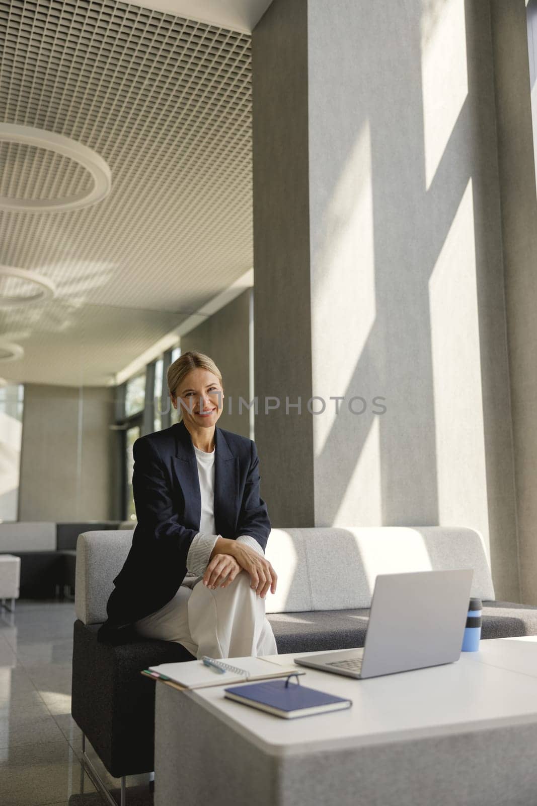 Smiling business woman working on laptop sitting on background of modern office and looks camera by Yaroslav_astakhov