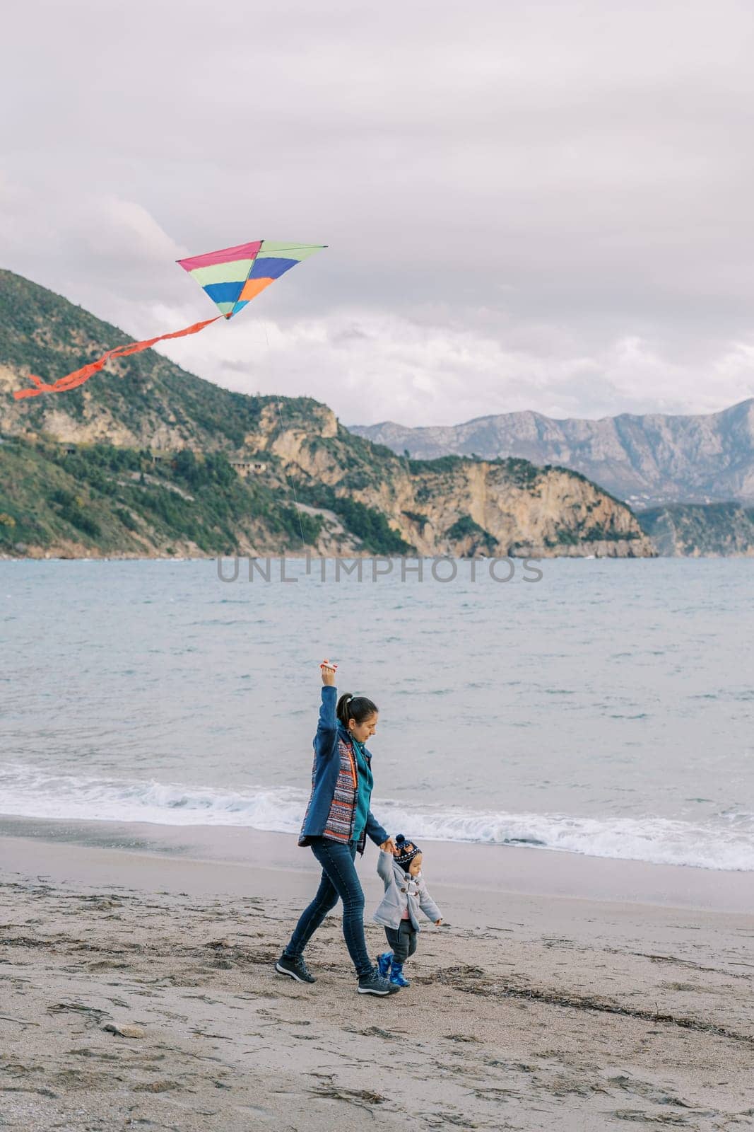 Mom with a little girl walk holding hands along the beach with a kite on a rope. Side view. High quality photo